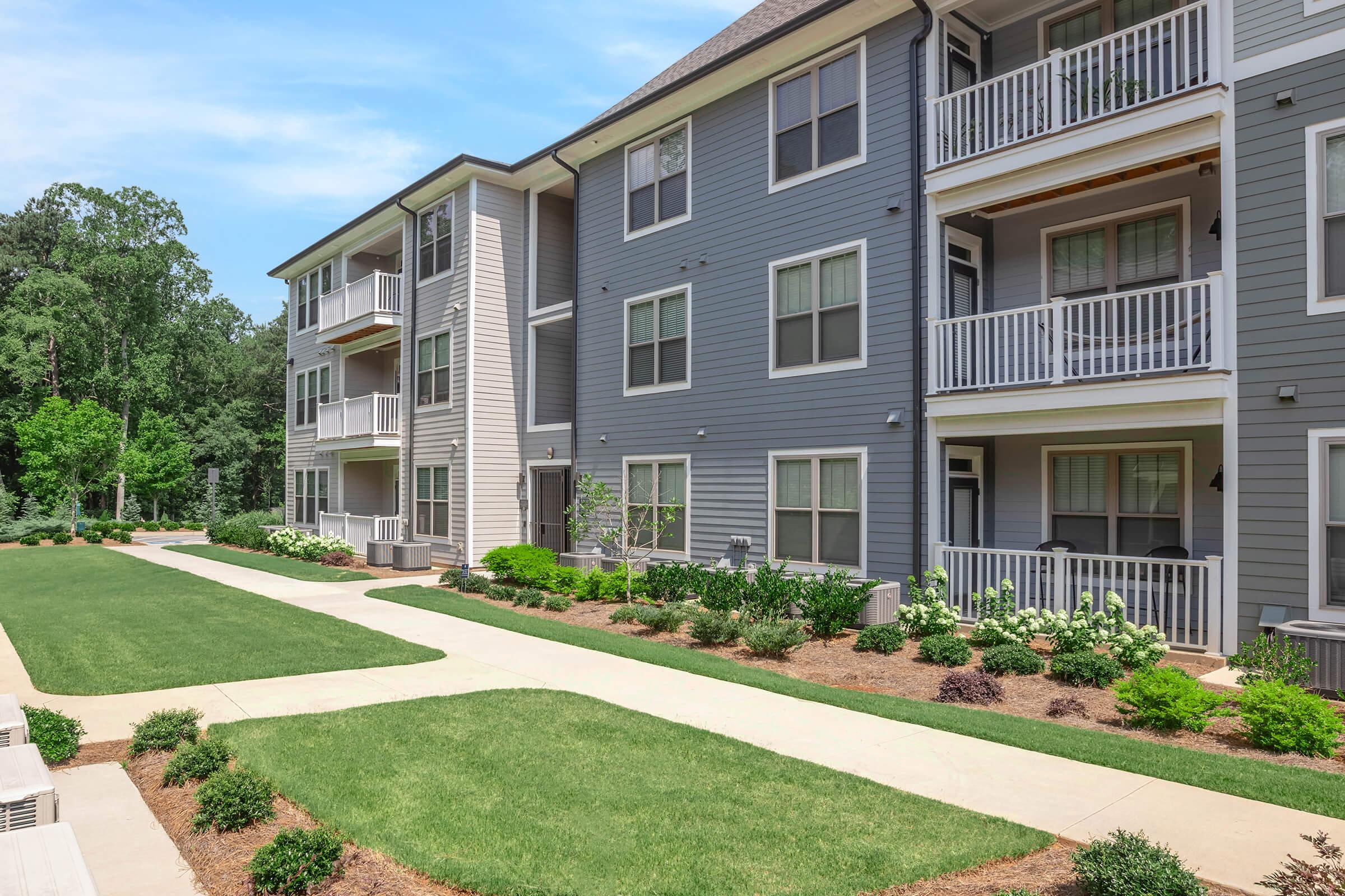 A modern apartment building featuring multiple stories with balconies, surrounded by well-maintained green lawns and landscaping. The pathway in front leads to the entrance, with bushes and flowers adding to the aesthetic appeal. The setting is bright and inviting, with clear blue skies in the background.