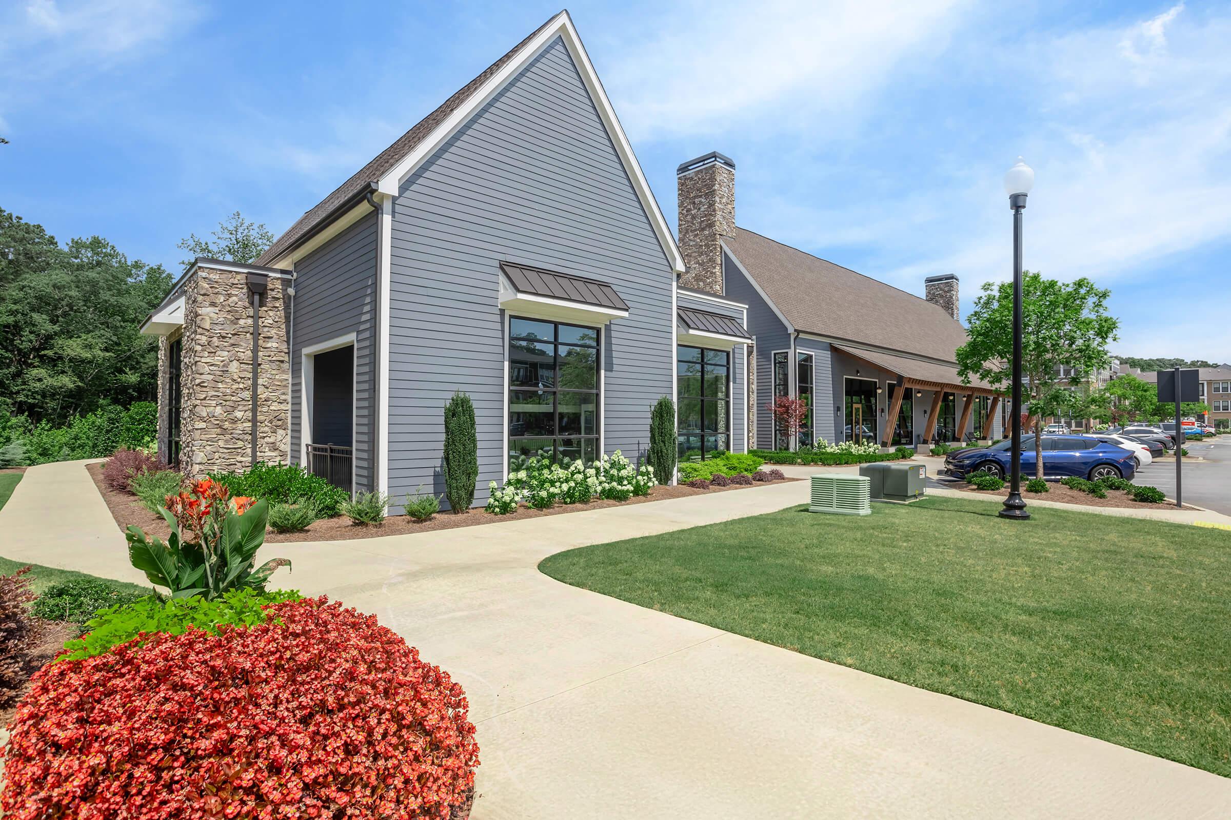 A modern building with a gray wooden exterior and stone accents, surrounded by neatly landscaped gardens featuring colorful flowers and shrubs. The pathway leads to the entrance, with parking spaces visible in the background and trees providing green foliage. A clear blue sky enhances the bright atmosphere.