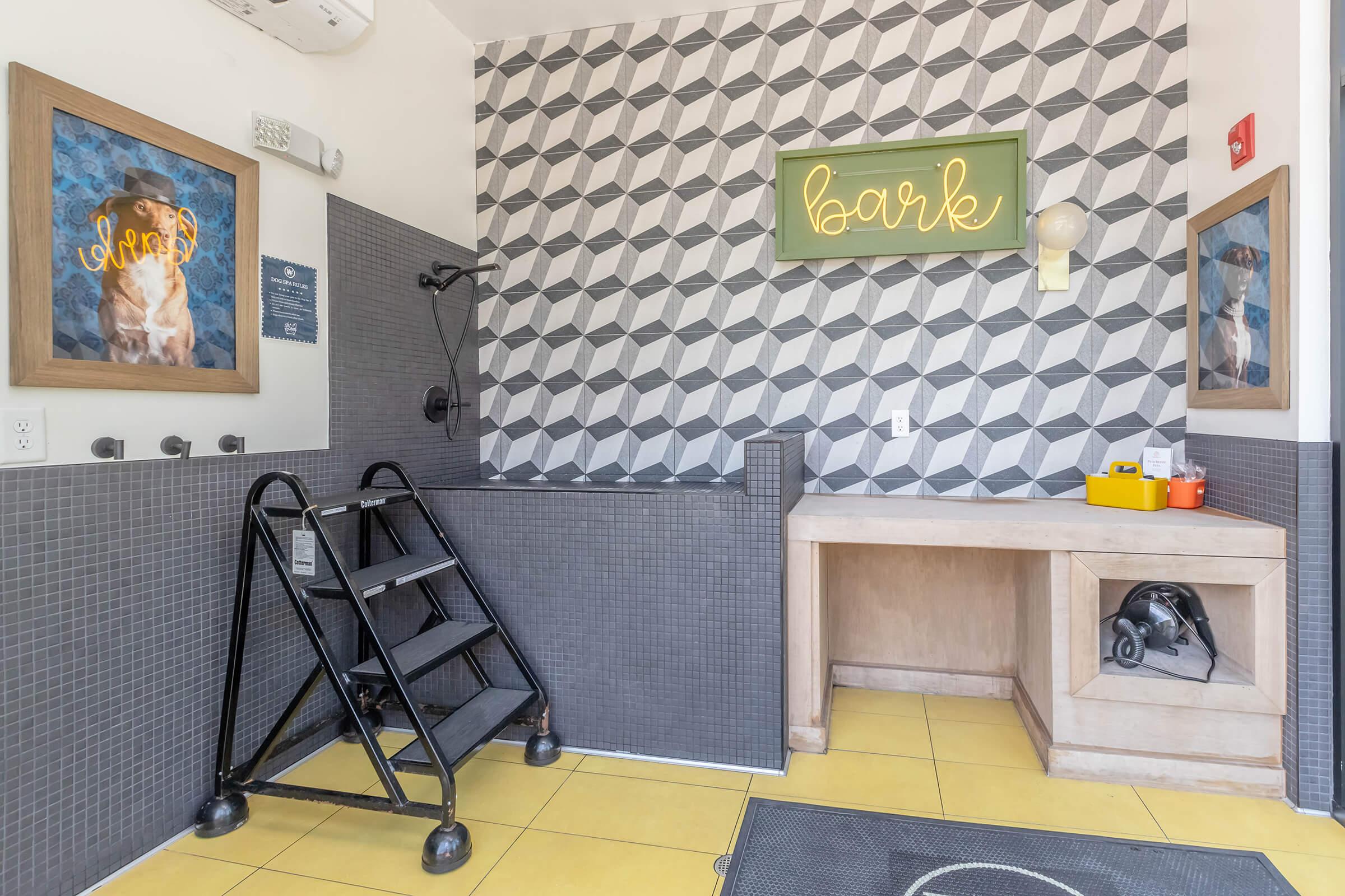 A spacious pet bathing area featuring gray tiled walls with geometric patterns, a shower area with a mounted hose, and a wooden table with grooming supplies. The word "bark" is illuminated in a green neon sign, creating a welcoming atmosphere for pet owners.