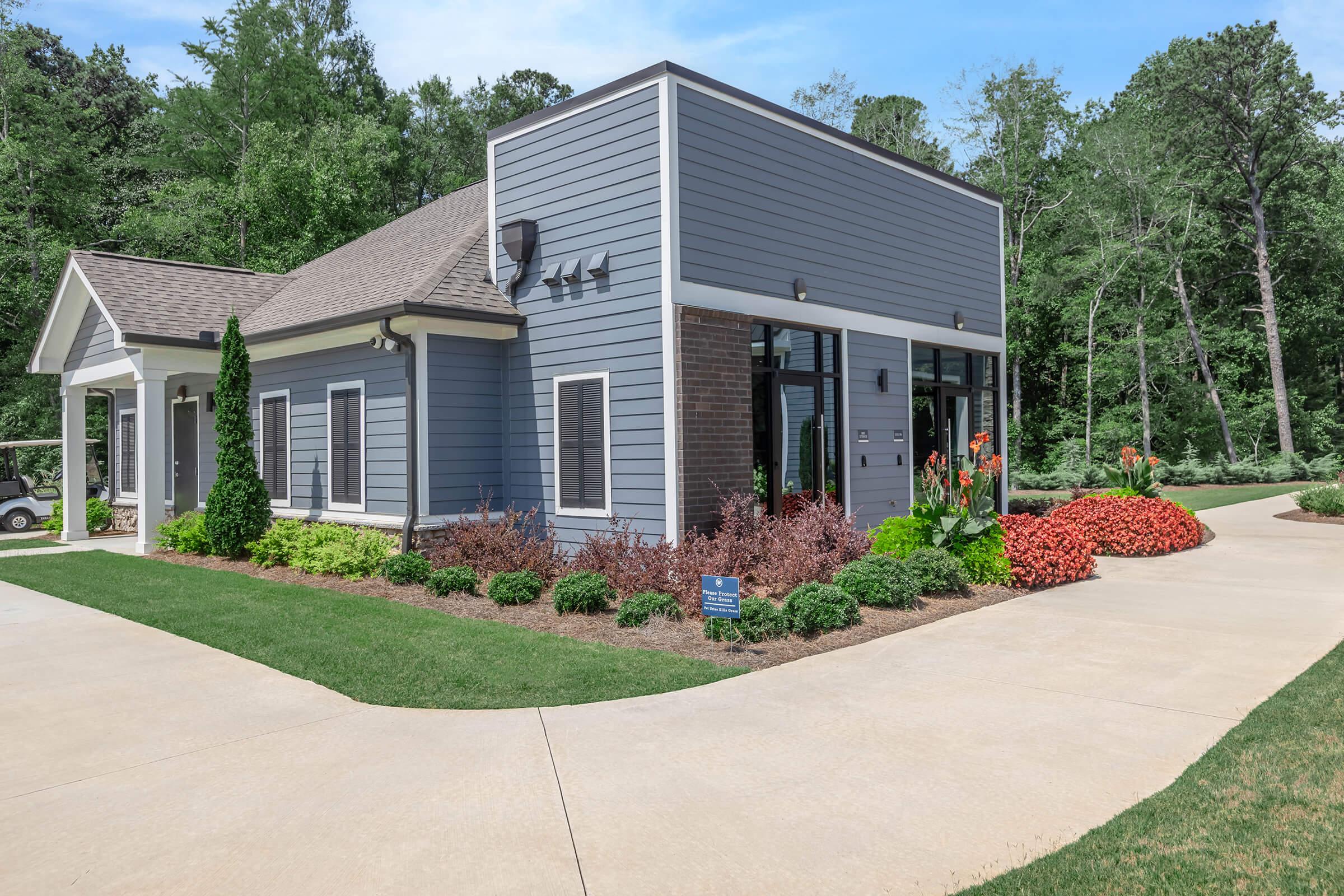 Single-story building with modern grey siding and brick accents, surrounded by well-maintained landscaping featuring colorful flower beds and green shrubbery. A concrete pathway leads to the entrance, with trees visible in the background under a clear blue sky.