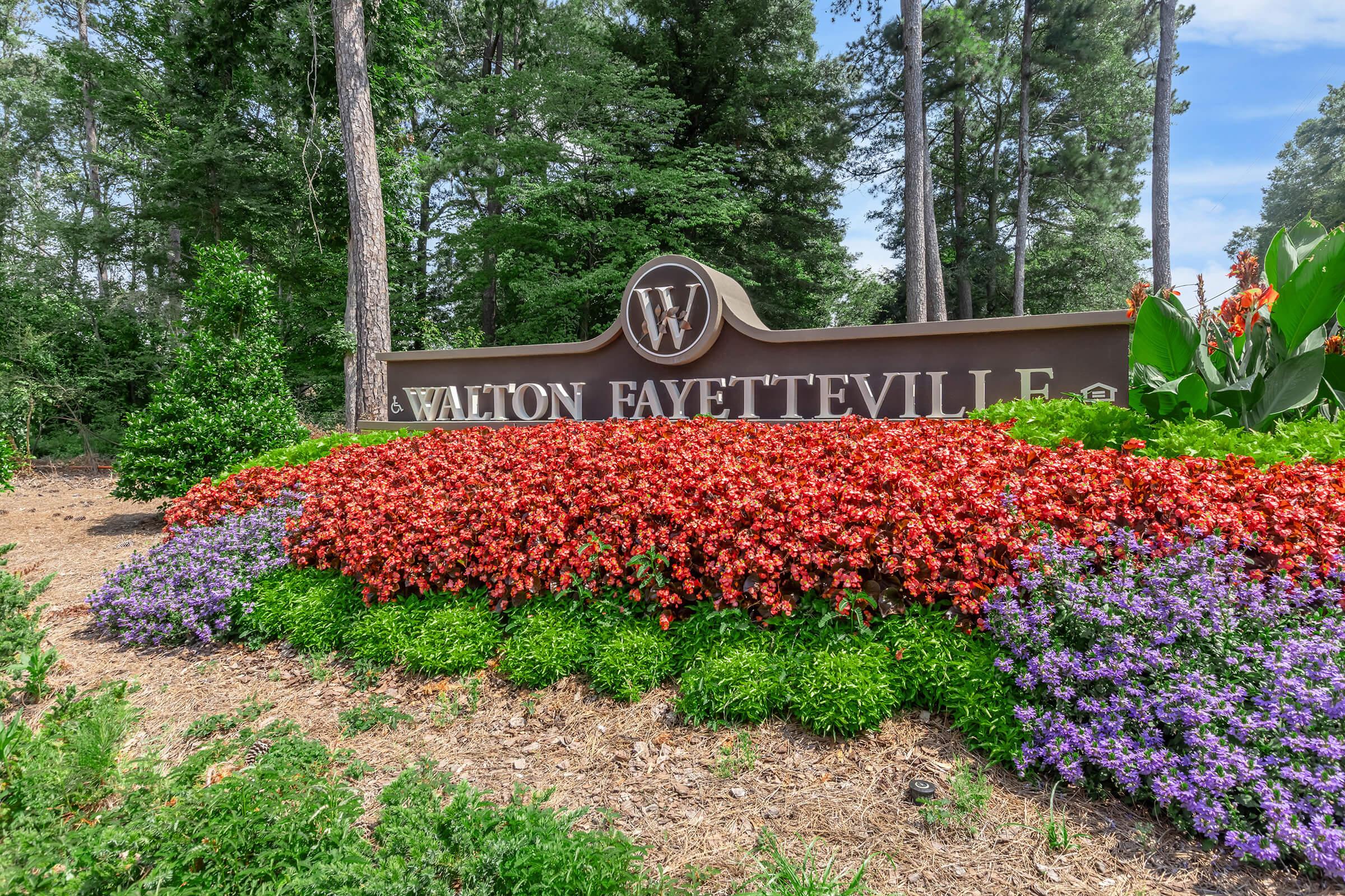 A decorative sign for Walton Fayetteville, surrounded by vibrant flower beds featuring red, orange, and purple blooms among lush green foliage, set against a backdrop of tall trees.