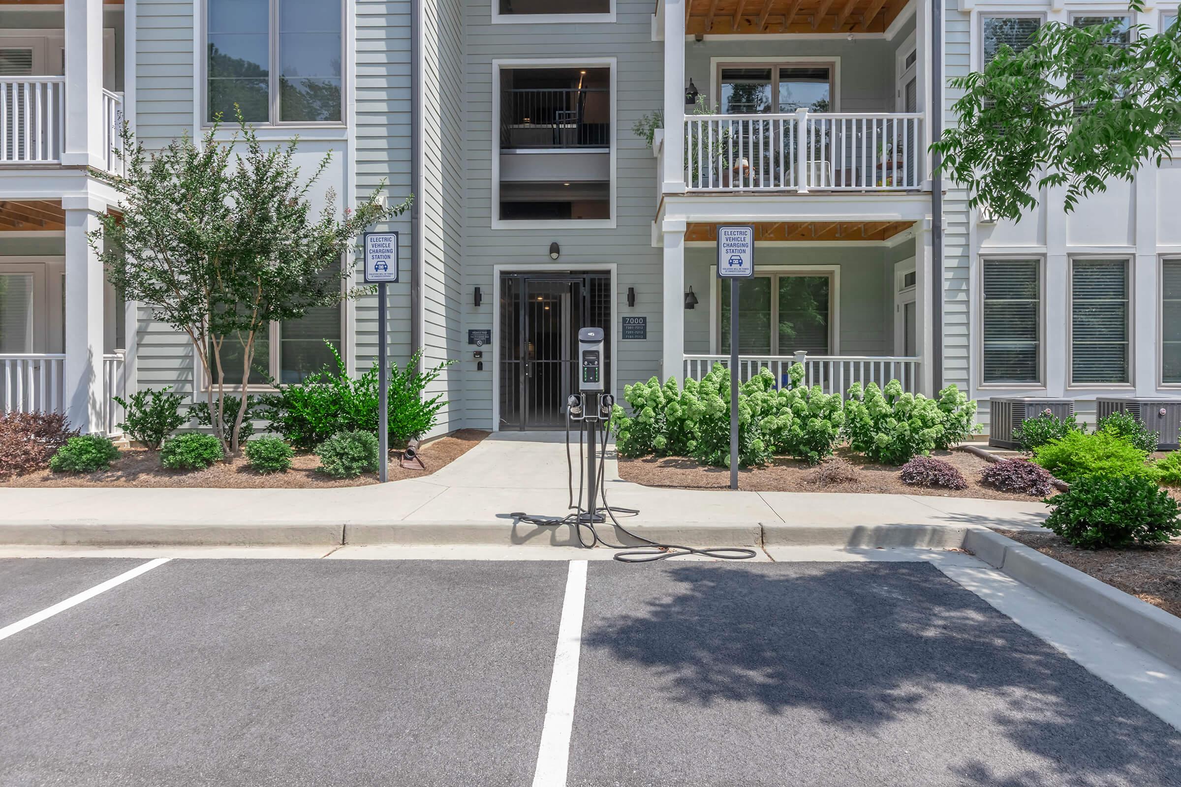 A modern apartment building entrance with multiple balconies, surrounded by landscaped greenery. In the foreground, there is a parking area with a designated space, along with a bike rack mounted on the sidewalk. The entrance is marked with signs indicating access points and rules.