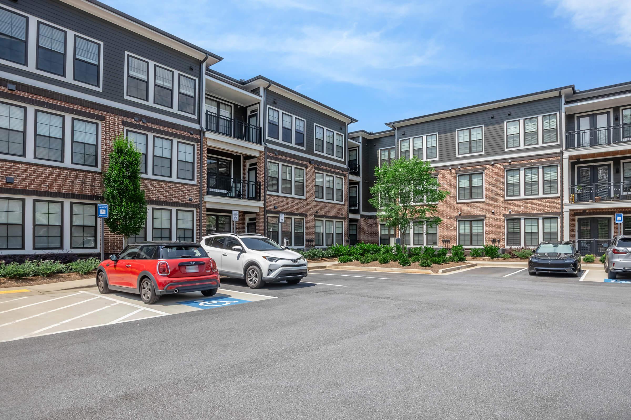 A view of a multi-story residential building with a combination of brick and modern siding. The parking lot features several cars parked in designated spaces, with some greenery and trees visible around the property. The sky is clear and blue, indicating a sunny day.