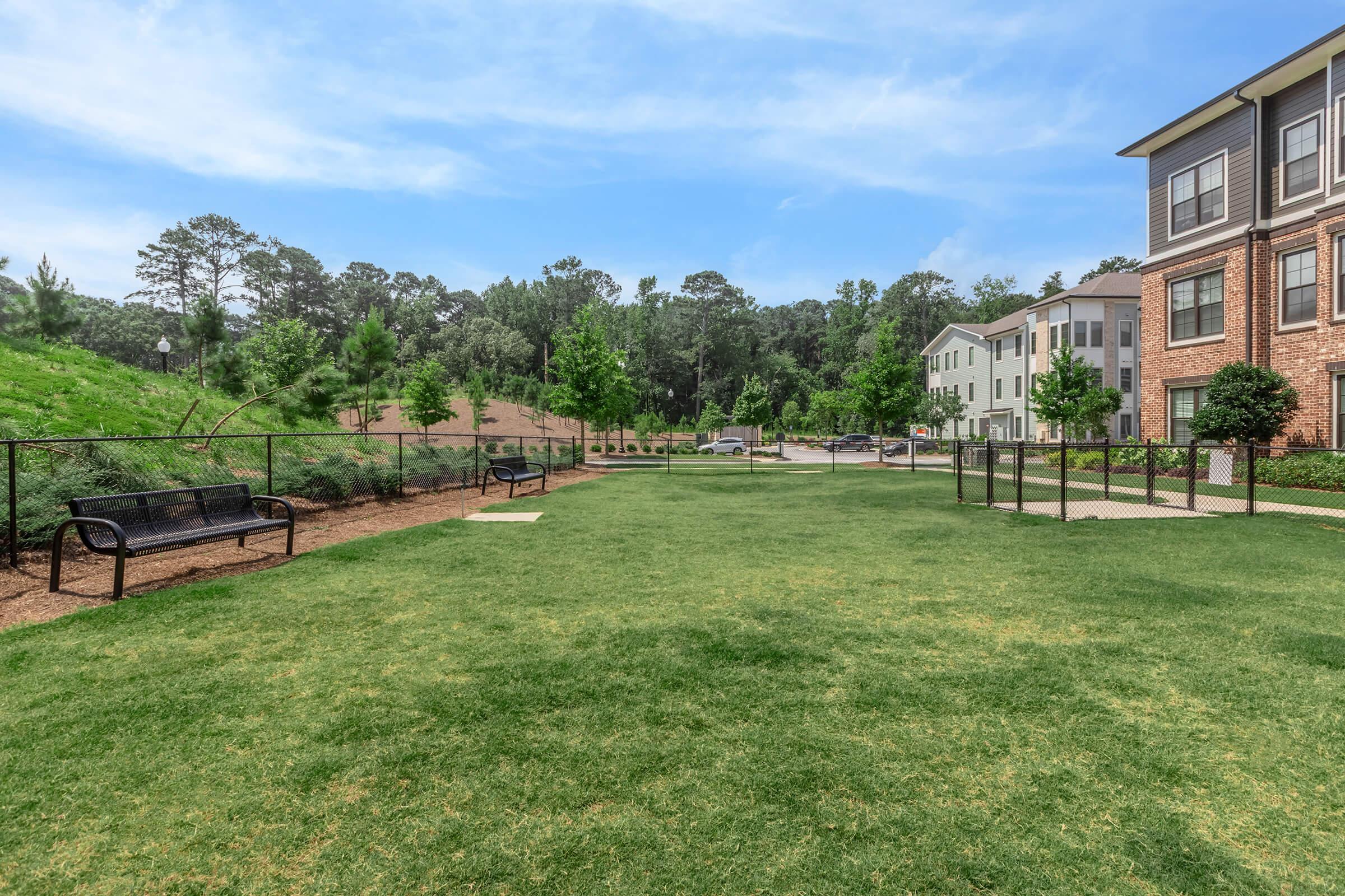 Green grassy area with benches and a black fence, surrounded by trees and shrubs. In the background, there are residential buildings and a pathway. The sky is blue with a few clouds, creating a serene outdoor atmosphere.