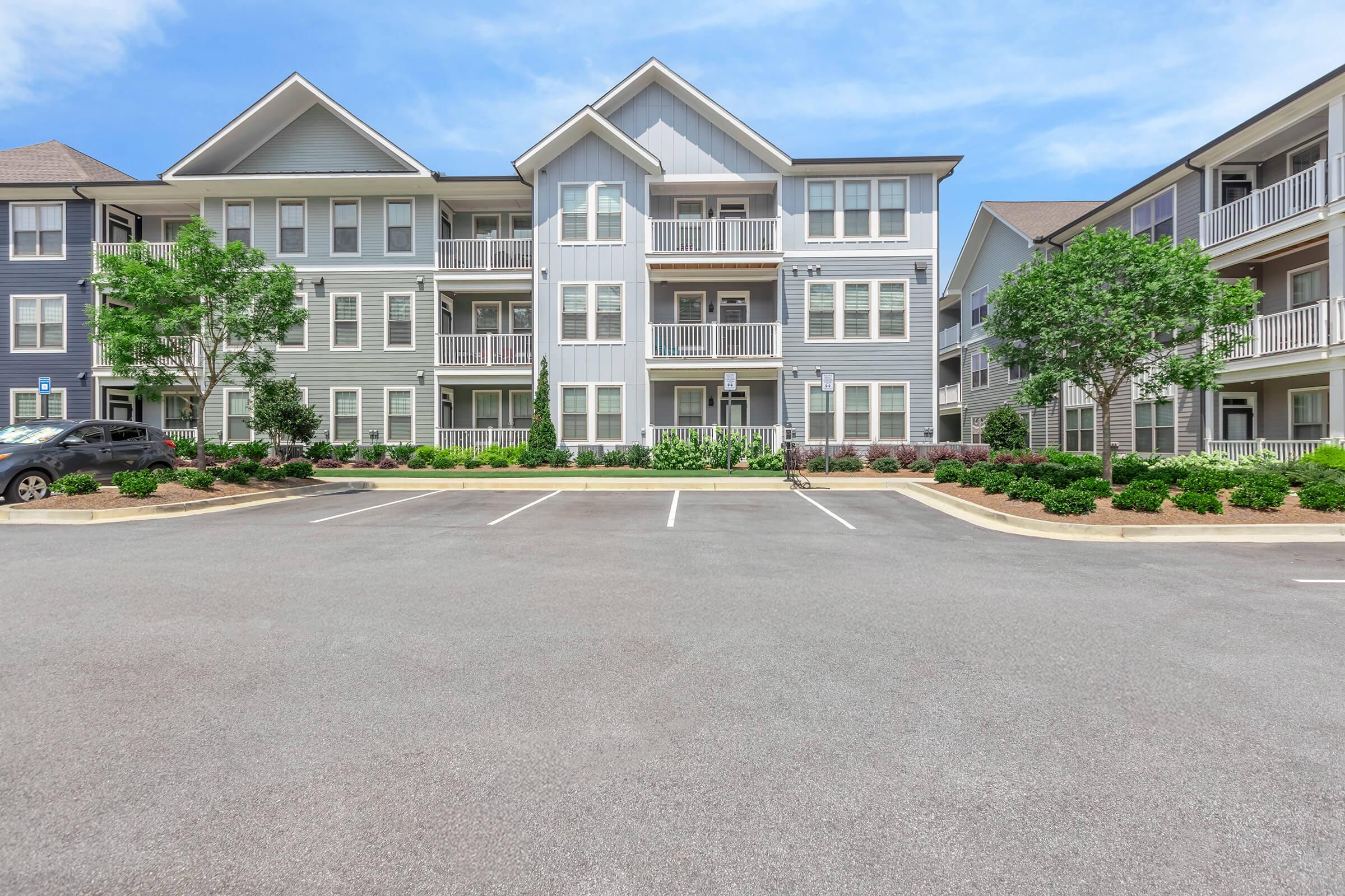 A modern apartment building with three floors, featuring a mix of wood and siding exteriors. The entrance is central with multiple balconies. Well-maintained landscaping includes shrubs and small trees. A parking area is visible in the foreground with several empty spaces. Clear blue sky in the background.