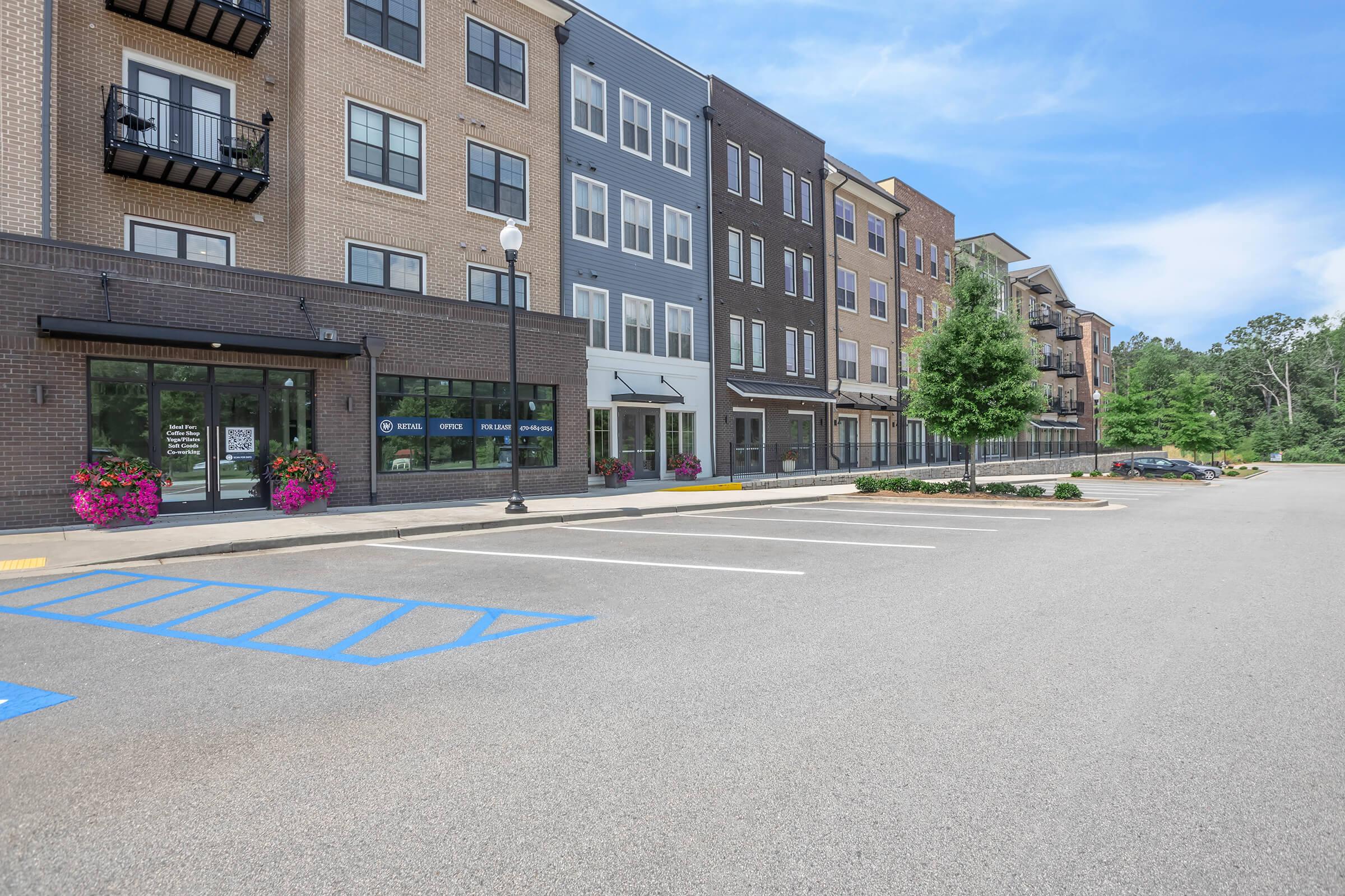 Modern apartment buildings with a mix of brick and siding exteriors. The foreground shows an empty parking lot with blue parking spaces. There are colorful flower planters near the entrance of the buildings. The background features more apartment units, with trees and a clear blue sky above.