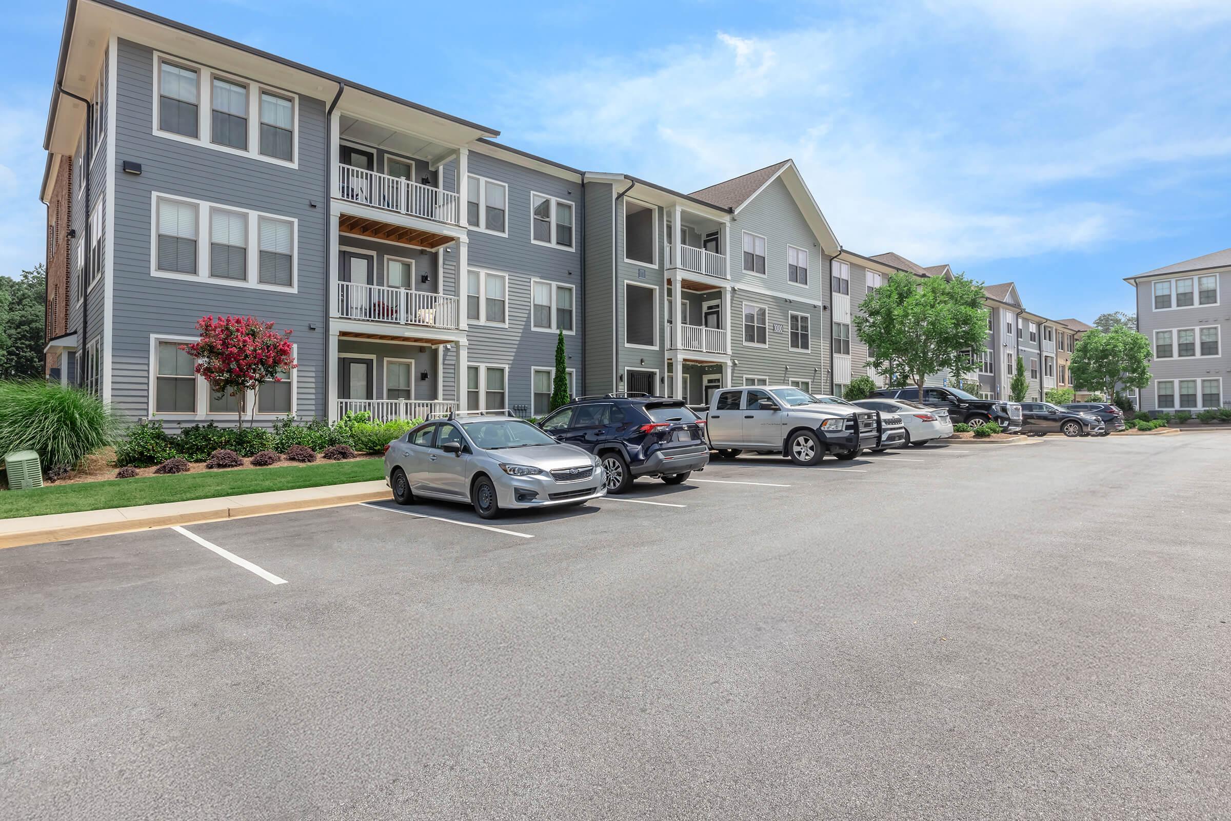 A view of a modern apartment complex with multiple gray buildings. In the foreground, there's a parking lot with several parked cars. Lush greenery and flowerbeds line the pathways, creating a welcoming environment. The sky is blue with a few clouds, indicating a clear day.