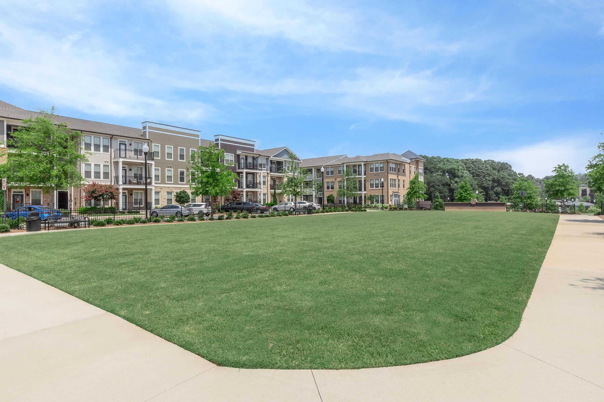 A well-maintained grassy area surrounded by modern apartment buildings. The scene features trees and a clear blue sky, providing a tranquil outdoor space. Paved pathways meander through the area, suggesting a peaceful community setting ideal for leisure activities.