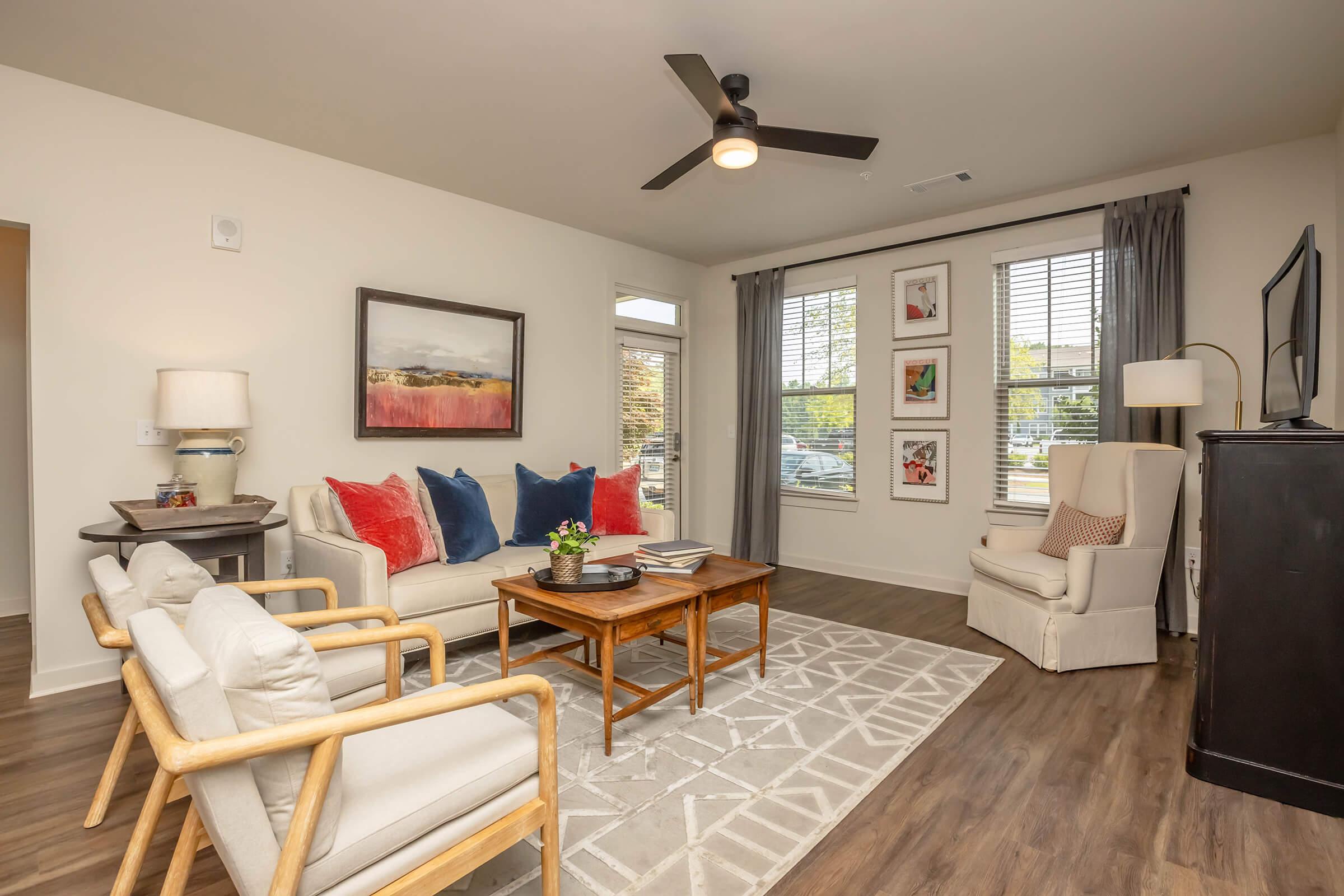 A modern living room featuring a neutral color palette. There is a light-colored sofa adorned with red and blue pillows, two wooden armchairs, and a wooden coffee table. The room includes framed artwork on the walls, a ceiling fan, a television, and large windows with curtains allowing natural light to illuminate the space.
