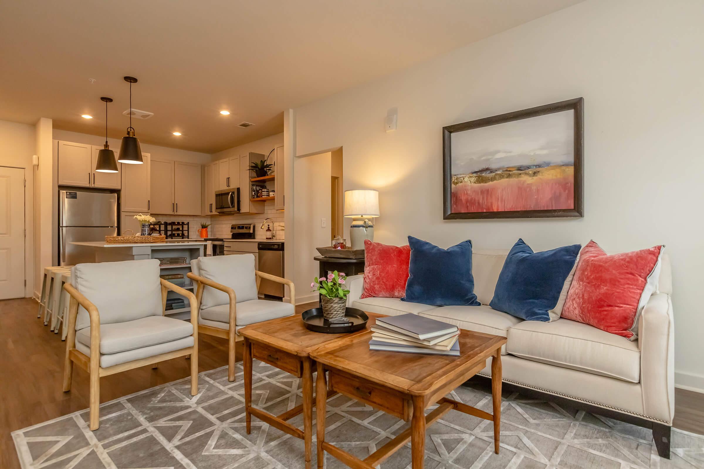 A modern living room featuring a light-colored sofa with colorful cushions, a wooden coffee table, and a potted plant. The open space includes a kitchen with stainless steel appliances in the background, along with a framed painting on the wall and stylish pendant lighting.
