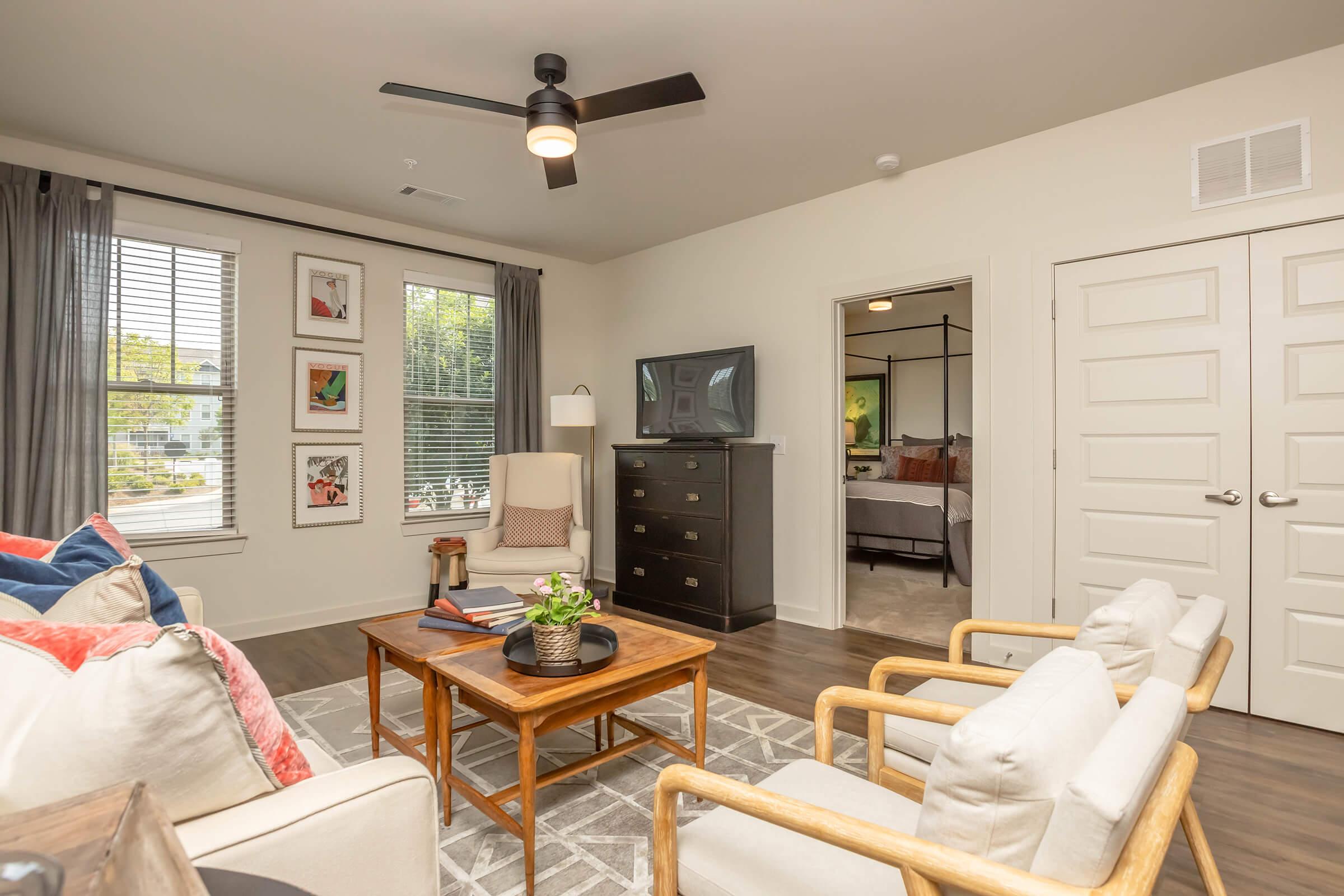 A cozy living room featuring a light-colored sofa with colorful throw pillows, a wooden coffee table, and two armchairs. A television is placed on a black dresser against the wall, while large windows allow natural light to brighten the space. The decor is modern with neutral tones and soft textures.