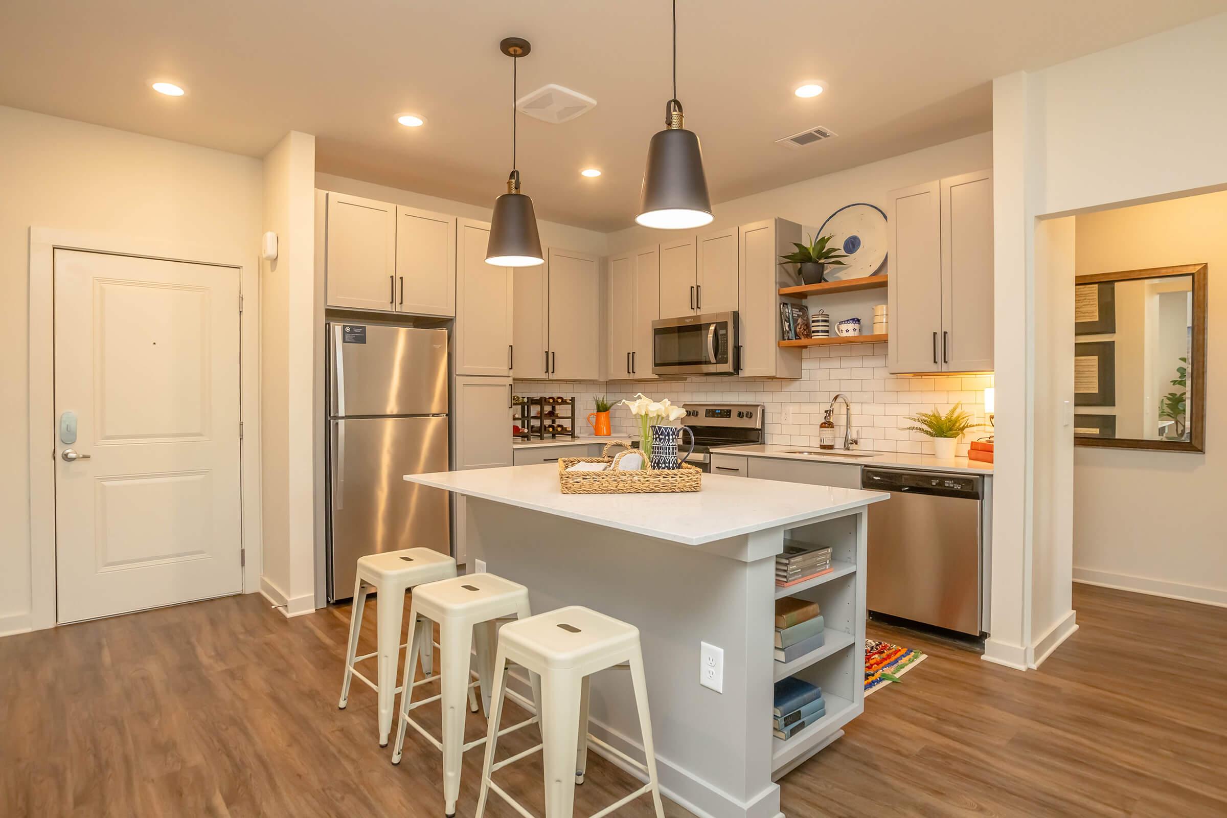 A modern kitchen featuring gray cabinetry, stainless steel appliances, and a white countertop island with three stools. The space is well-lit with pendant lights and has a stylish decor, including a decorative plate and fresh flowers. The walls are light, and the flooring is a warm wood tone.