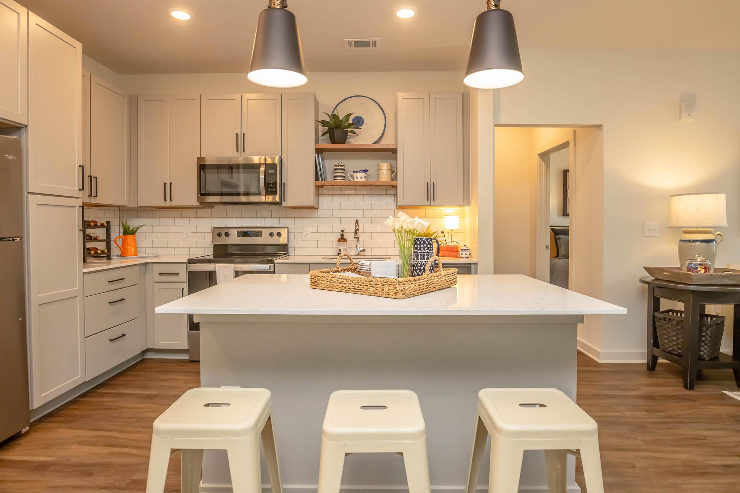 Modern kitchen featuring light grey cabinets, stainless steel appliances, and a white quartz island with three metal stools. Pendant lights hang over the island, and decorative items are displayed on shelves. The space is well-lit, creating a warm and inviting atmosphere.