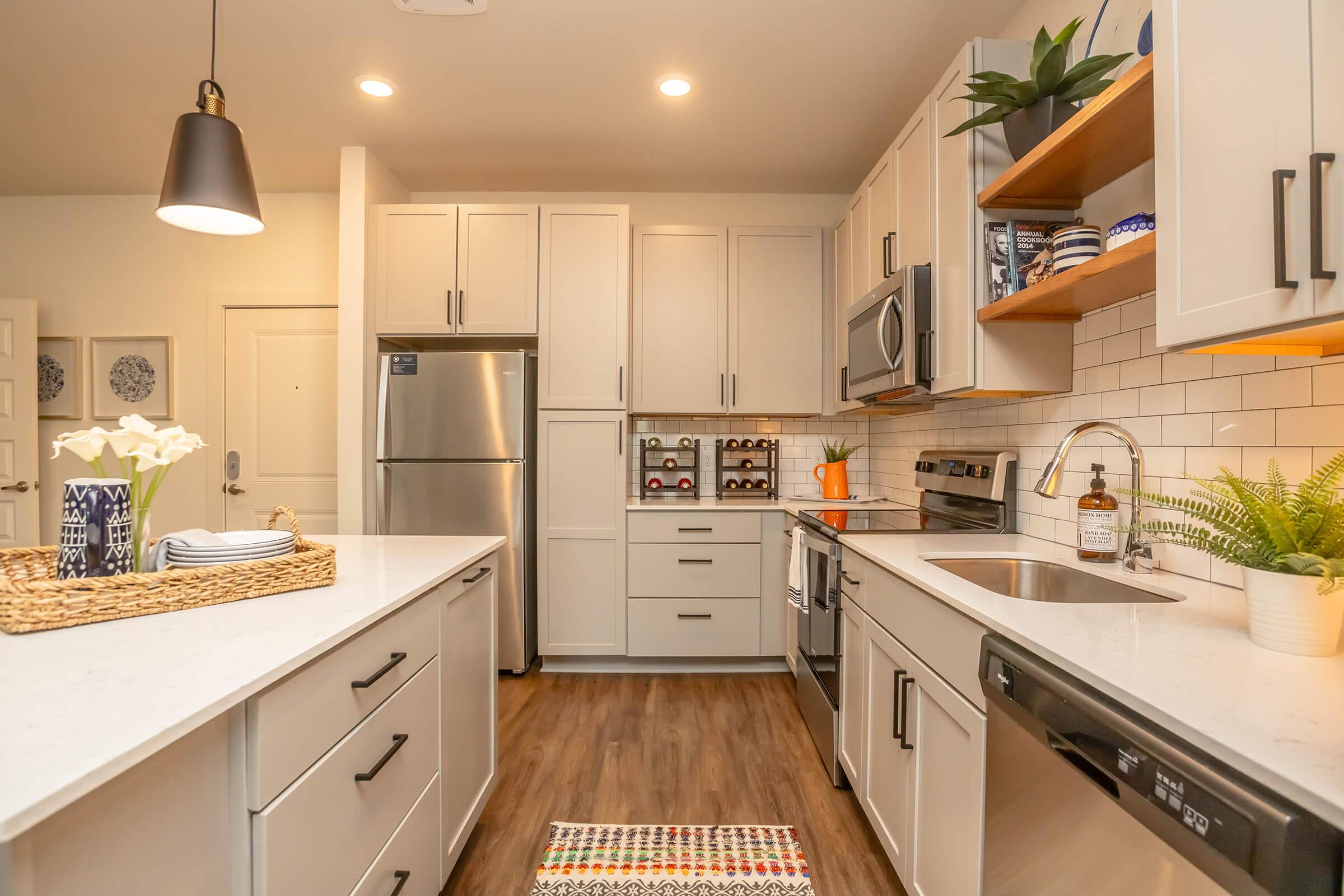 Modern kitchen featuring a clean design with white cabinets, stainless steel appliances, and a breakfast bar. There are open shelves displaying dishware, a wine rack, and a cozy area rug. Bright lighting and green plants add warmth to the space.