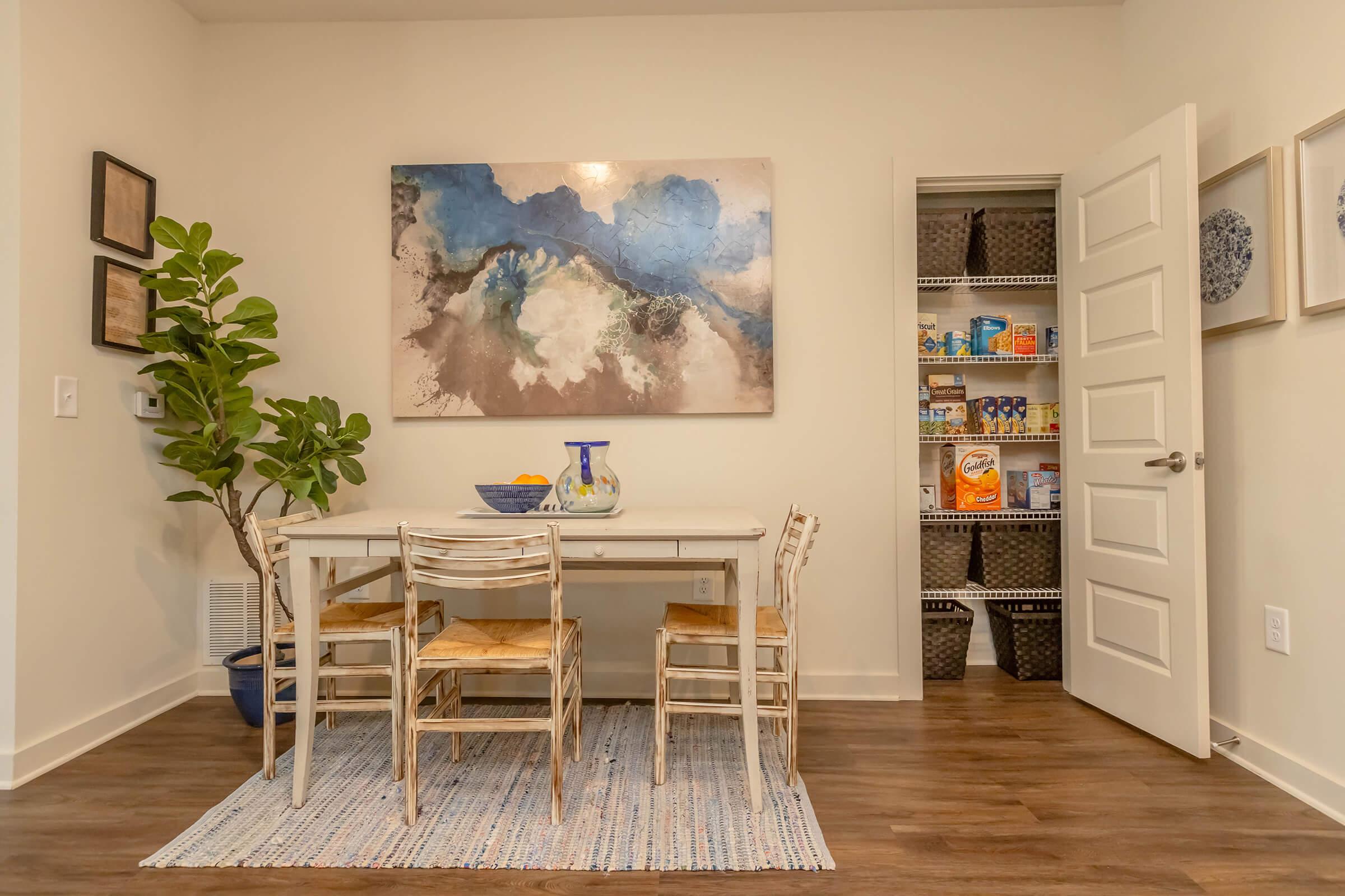 A bright dining area featuring a wooden table surrounded by four chairs, a large abstract painting on the wall, a green plant in a pot, and a pantry door slightly ajar showcasing organized shelves with various food items and containers. The floor is covered with a textured rug.