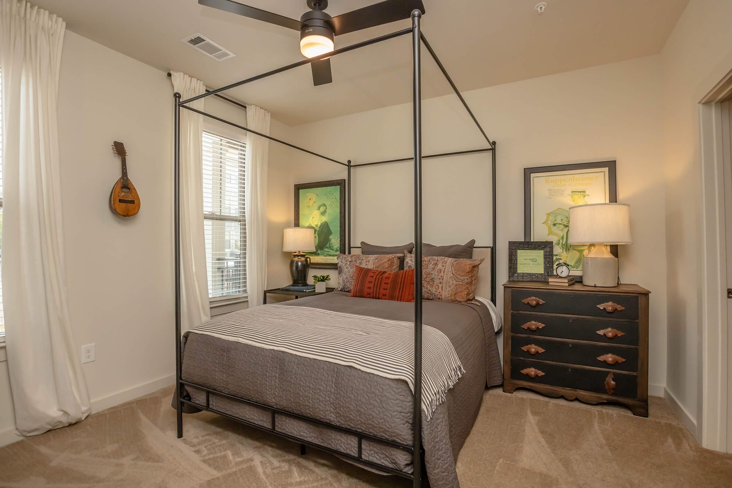 A cozy bedroom featuring a black four-poster bed with decorative pillows, two bedside lamps, and a dark wood dresser. There are soft beige curtains and a guitar hanging on the wall. Two framed artworks are displayed above the bed, adding artistic flair to the space. The room has warm, inviting tones and plush carpeting.