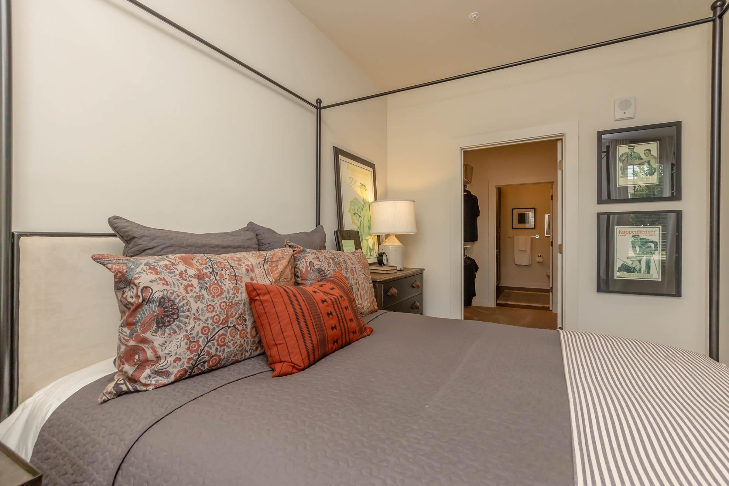 A cozy bedroom featuring a four-poster bed with decorative pillows in shades of gray and orange. A bedside table with a lamp is positioned next to the bed. The walls are adorned with framed art pieces, and a bathroom doorway is visible in the background, adding to the room's inviting atmosphere.