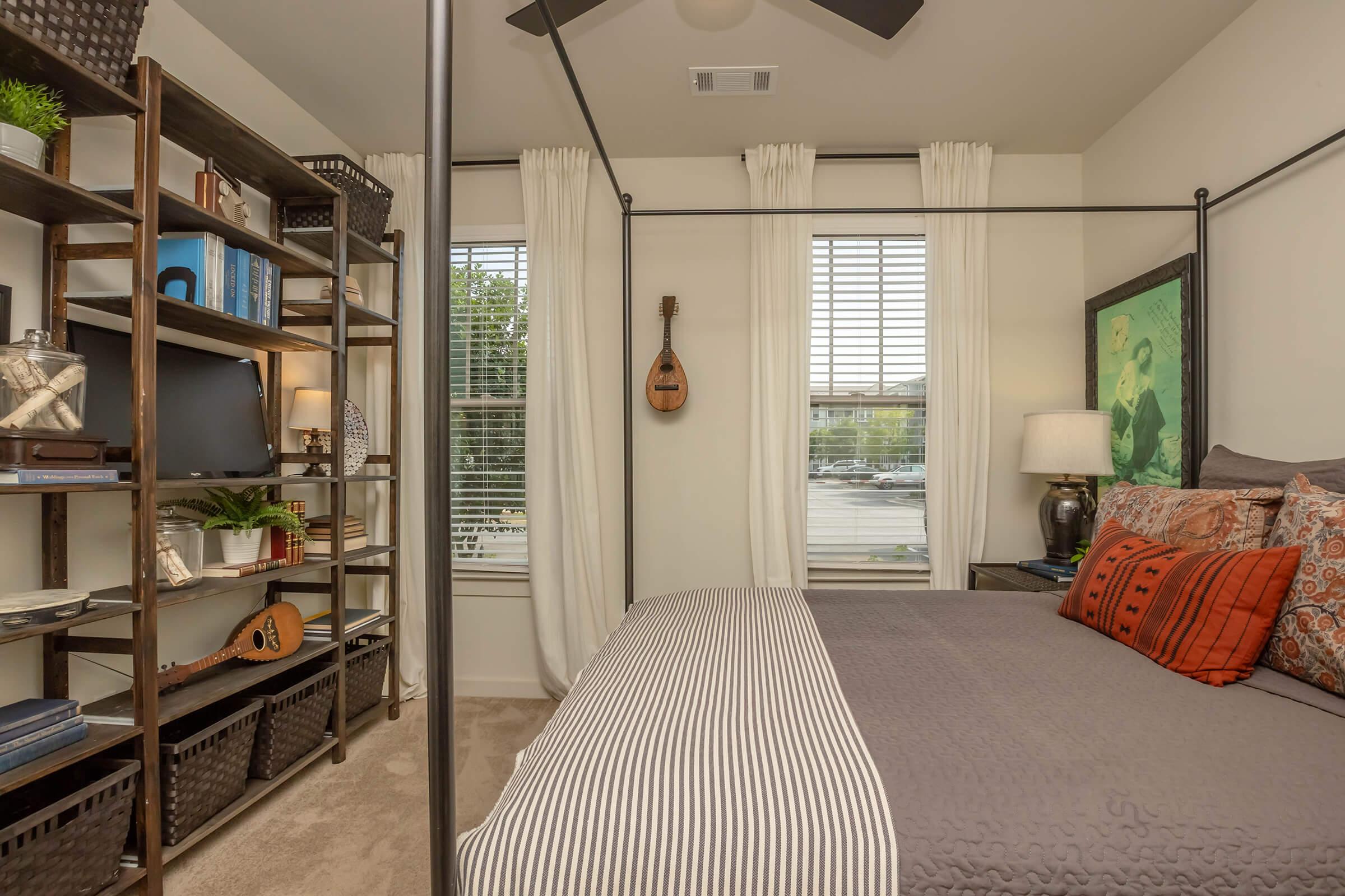 A cozy bedroom featuring a four-poster bed with decorative pillows, a bedside lamp, and a large window with sheer curtains. A stylish bookshelf with plants and decor is on one side, while a mandolin hangs on the wall. The room is bright and inviting, showcasing a blend of comfort and modern design.