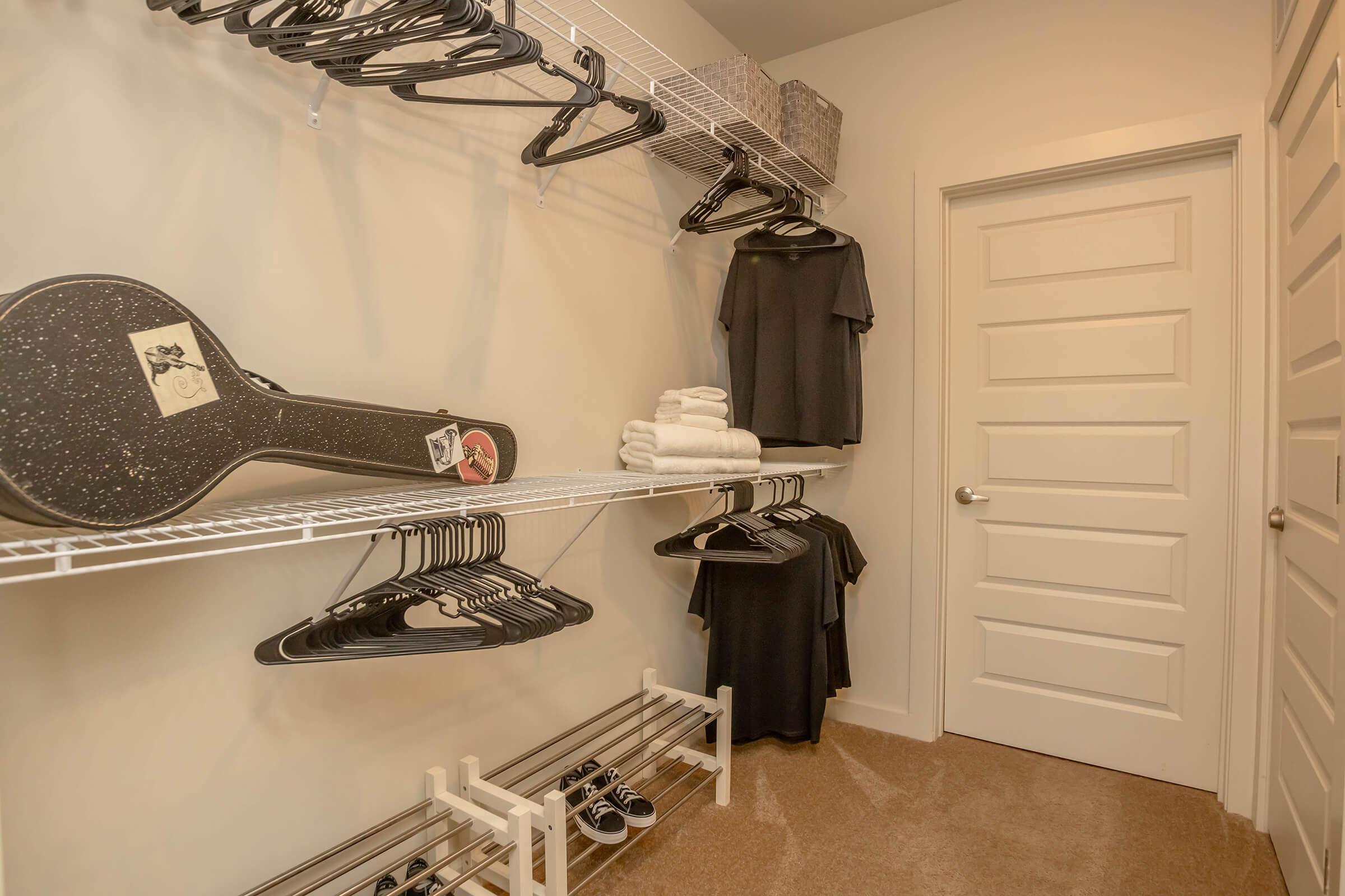 A neatly organized closet featuring a wire shelving unit with black hangers, two black shirts hanging, and a guitar case on the floor. There are also folded towels stacked neatly and a pair of shoes on a shoe rack. The closet has a door in the background, and the walls are painted a light color.