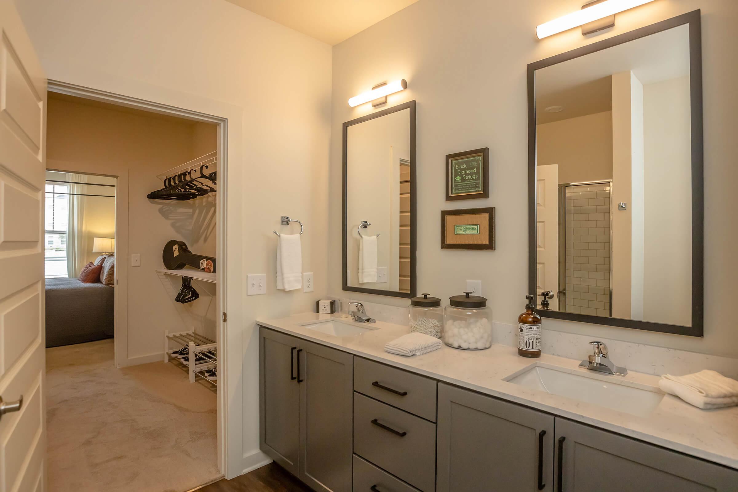 A modern bathroom with double sinks and rectangular mirrors above. The cabinetry is dark gray, and there's a countertop with neatly organized toiletries. To the side, an open door leads to a bedroom featuring a bed and a closet with clothes hangers. The overall design is sleek and contemporary.