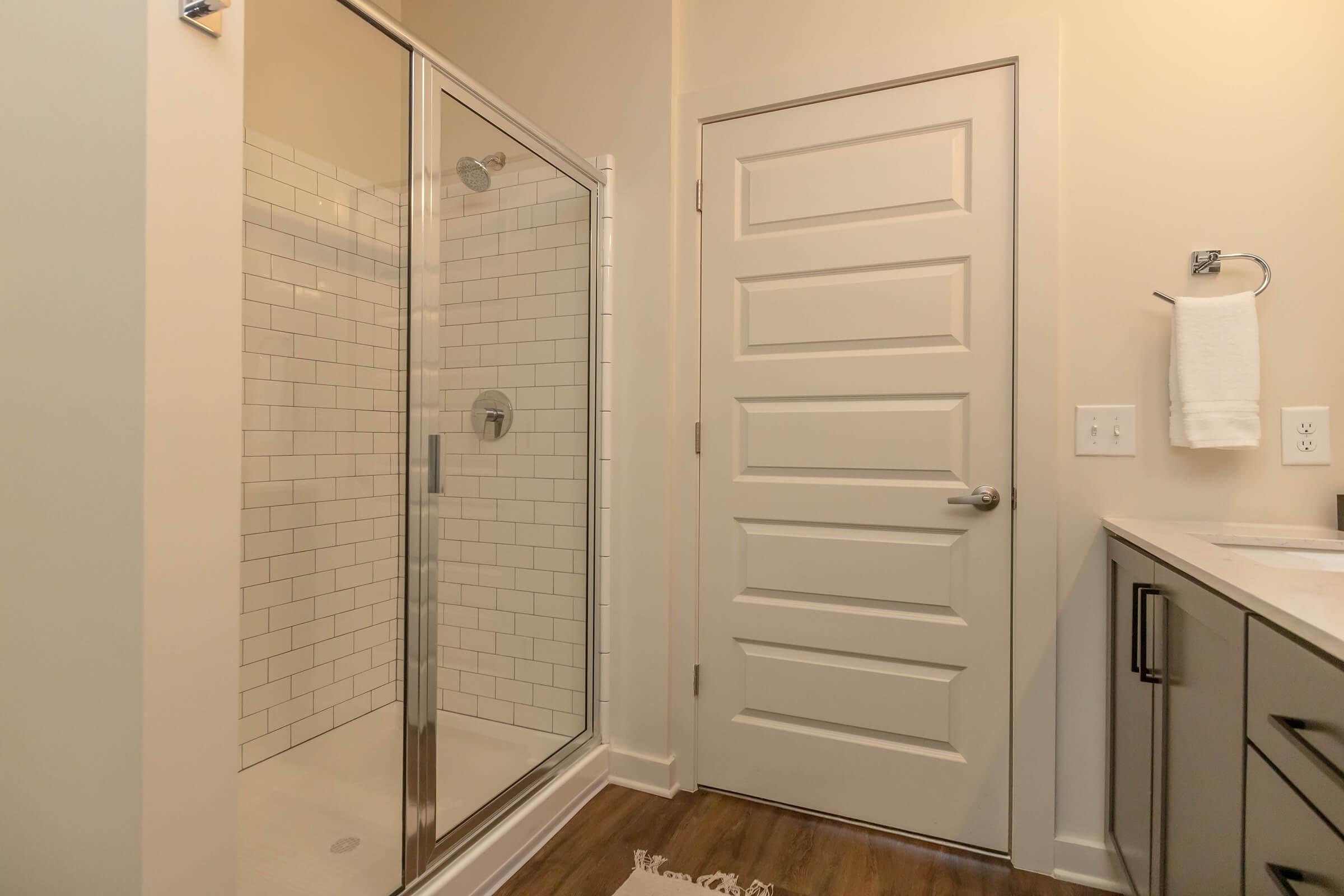 Modern bathroom featuring a glass shower enclosure with white subway tiles, a sleek vanity with dark cabinetry, a closed door, and a towel hanging on a rack. The room has light-colored walls and a wood-look floor, creating a clean and contemporary appearance.