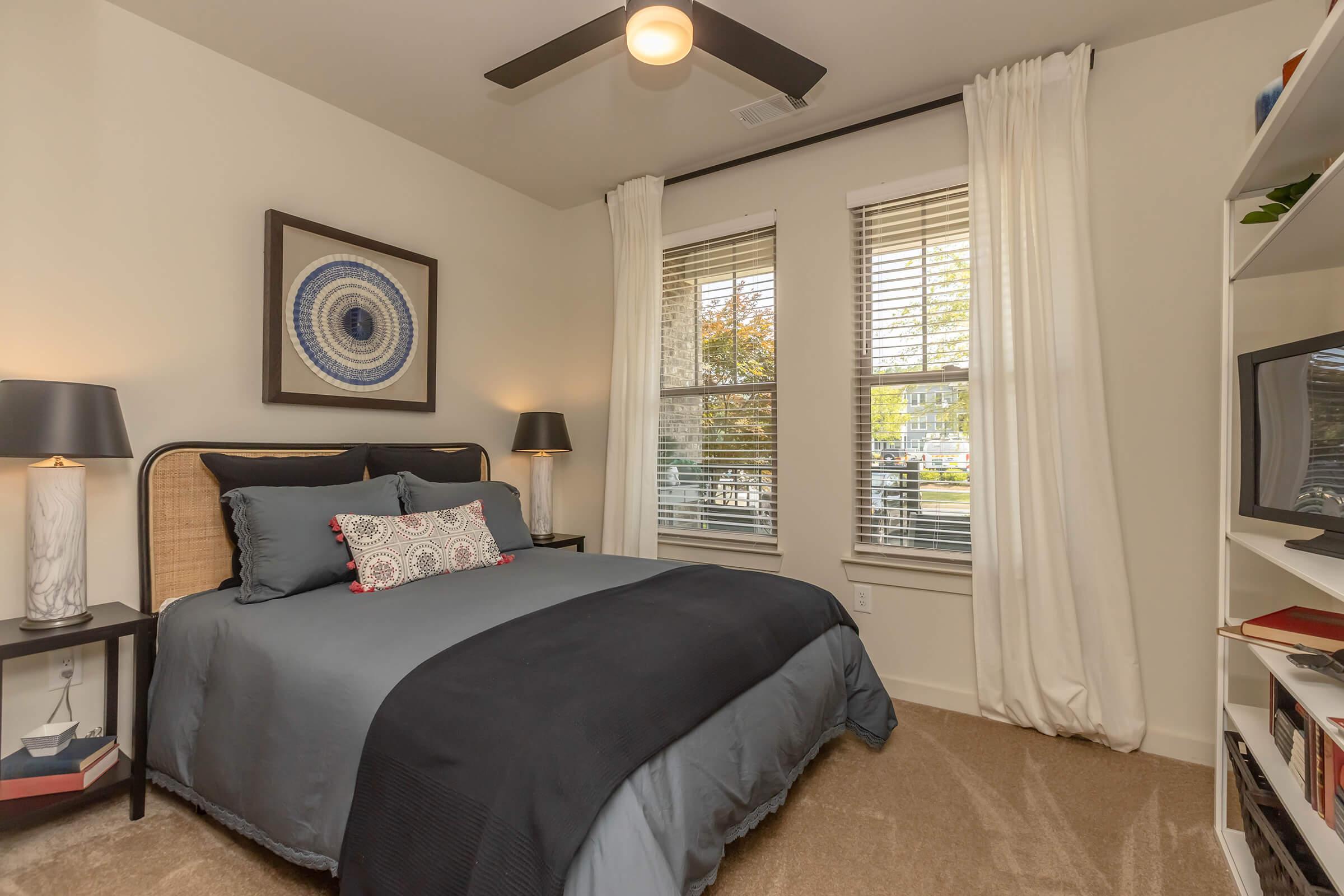 Cozy bedroom featuring a double bed with gray bedding, two bedside lamps, and decorative pillows. A framed circular artwork hangs above the bed. The room has large windows with sheer curtains, and a white bookshelf displaying books and decor. Warm, inviting atmosphere.