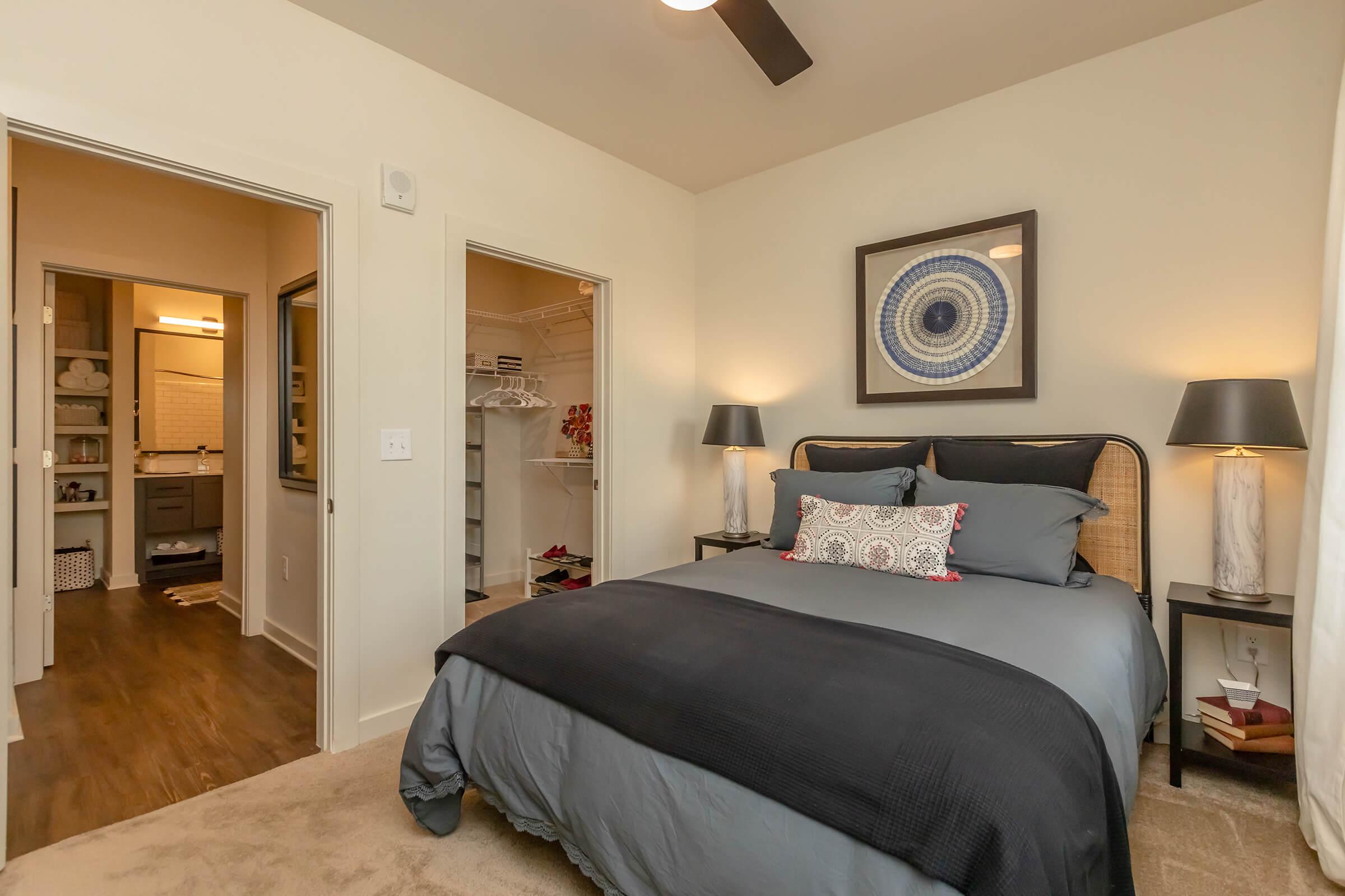 A cozy bedroom featuring a large bed with dark bedding, two bedside lamps, and a decorative wall art piece. To the side, there are doors leading to a closet and a bathroom. The room has a soft carpet and warm lighting, creating a comfortable and inviting atmosphere.