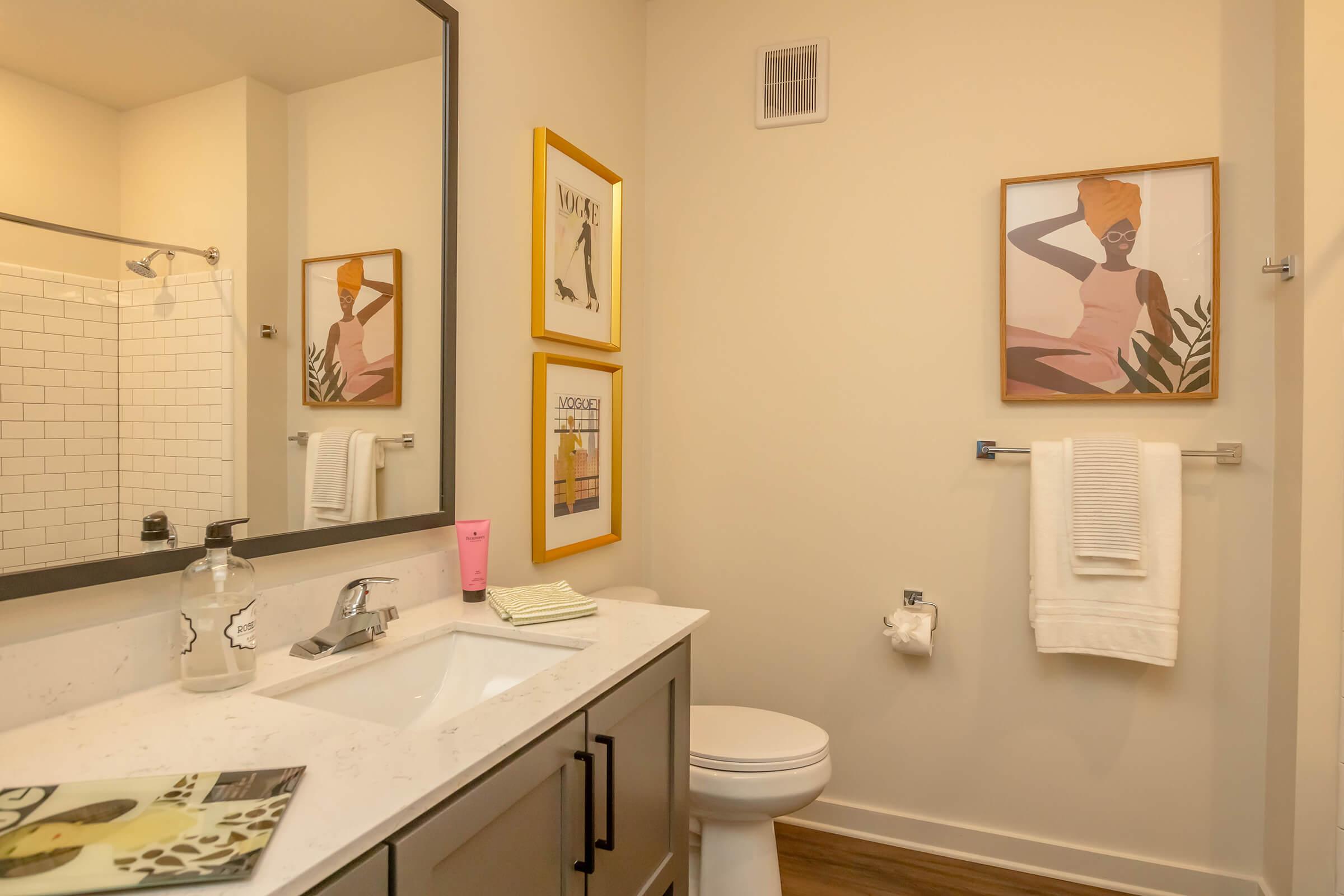 A modern bathroom featuring a white countertop sink with a mirror above, decorative wall art, and neatly arranged towels. There’s a shower with a glass door in the background, and the décor includes a plant and magazines on the counter, creating a clean and stylish atmosphere.