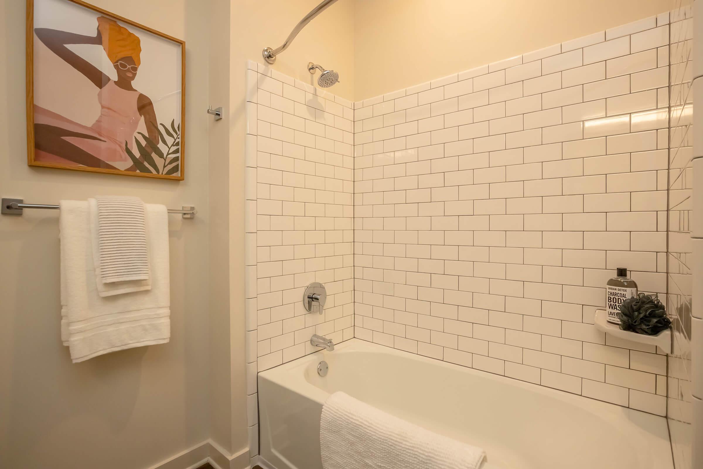 A modern bathroom featuring a white tiled bathtub with a shower, complemented by a framed artwork of a woman in a bathing suit. White towels hang neatly, and a decorative plant adds a touch of greenery to the space. The overall design is clean and stylish, with neutral tones and contemporary accents.