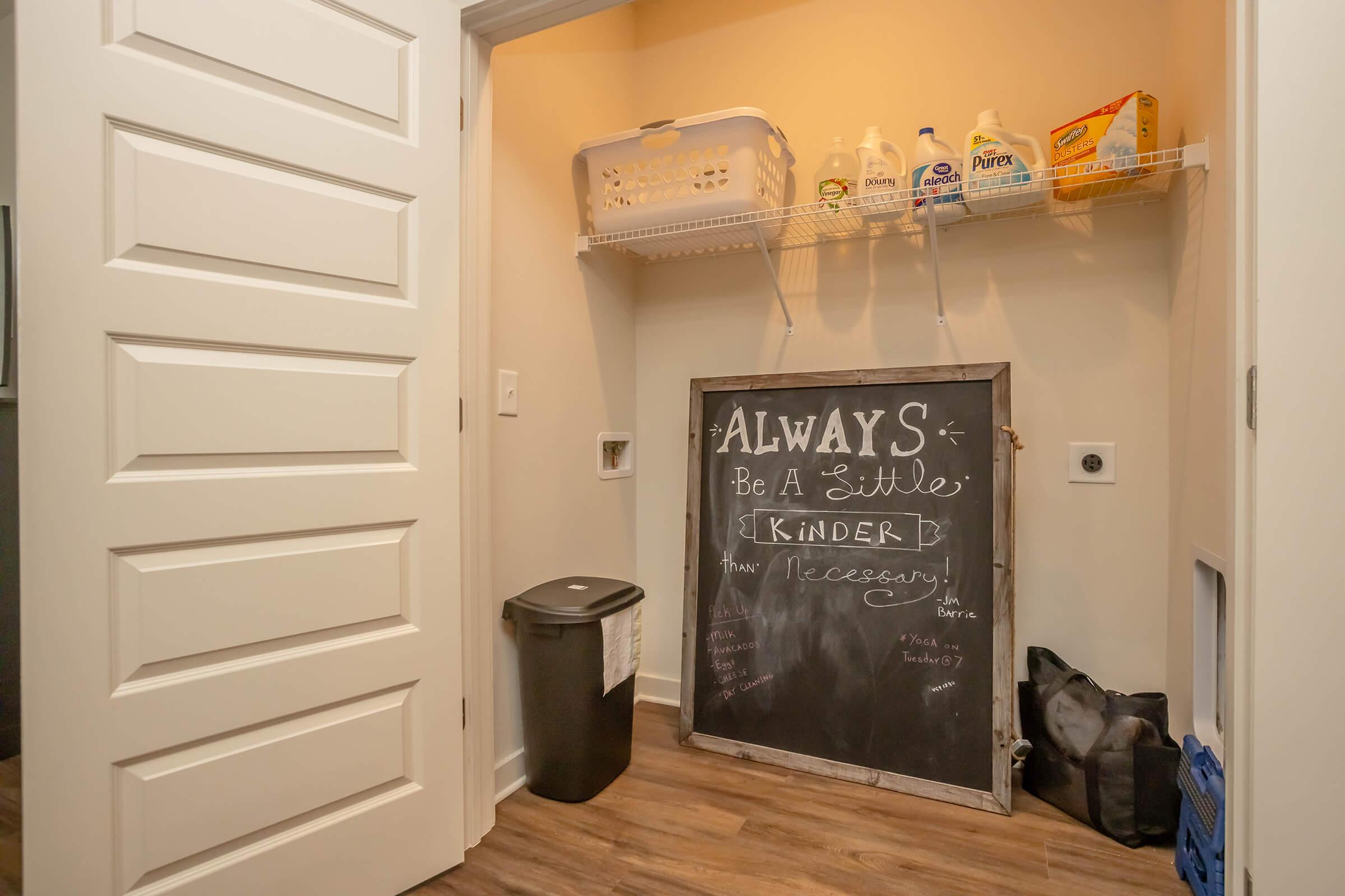 A small laundry room or closet with a white shelf holding various cleaning supplies. There's a chalkboard with the message "ALWAYS Be a Little Kinder" and a list of tasks or reminders. A trash can is visible, along with a laundry basket and a black bag on the floor. The walls are painted a light color.
