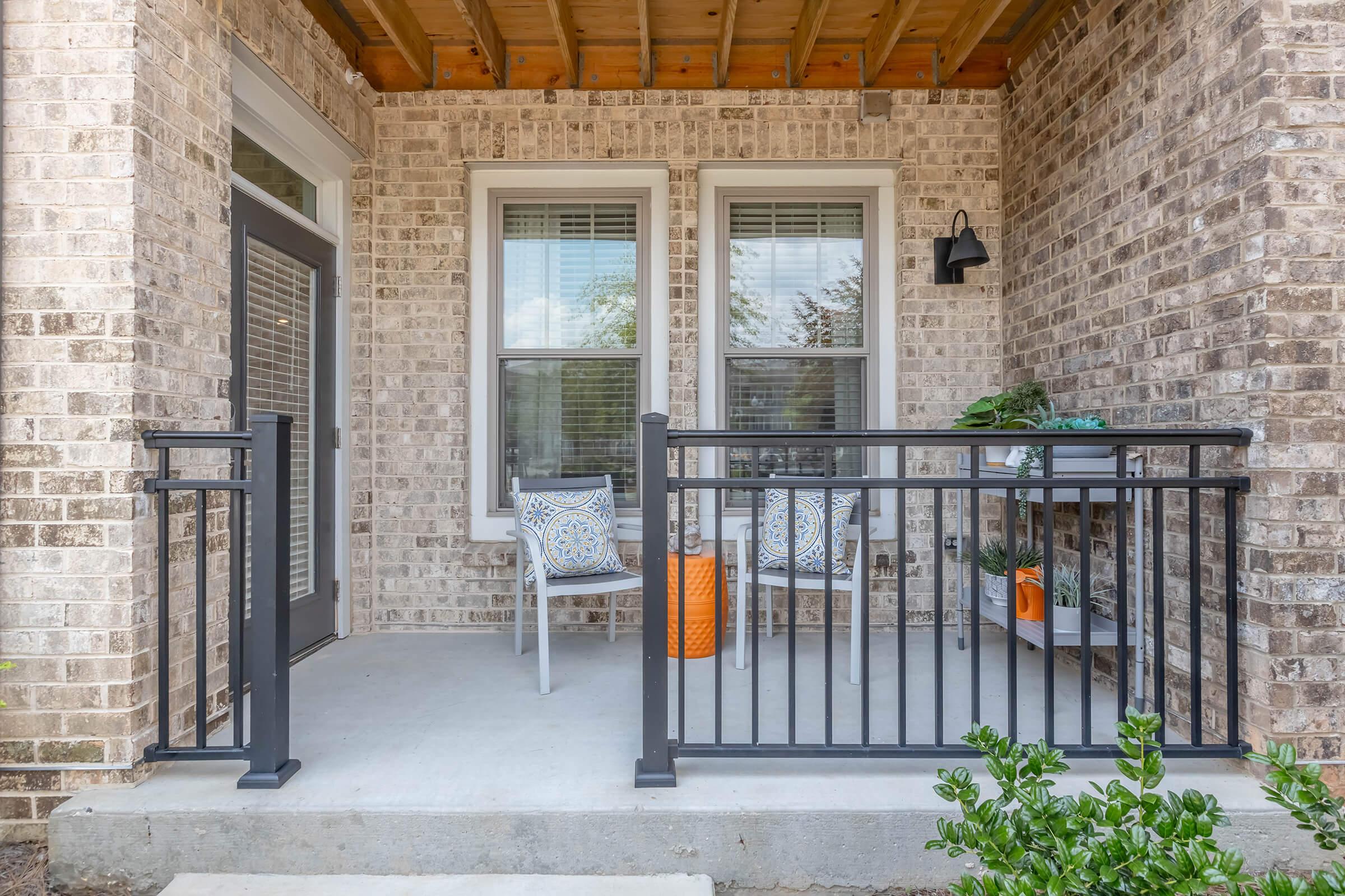 A welcoming front porch featuring a brick wall, two white chairs with decorative blue and orange cushions, and an orange side table. A small potted plant sits on the table. The space is partially enclosed by a black railing and has large windows allowing natural light.
