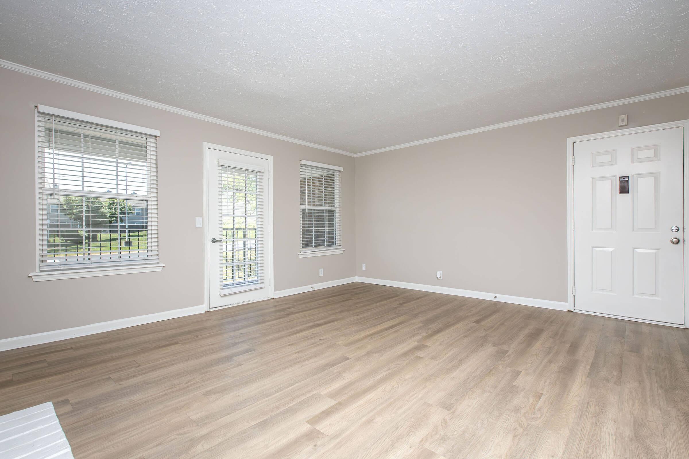 A spacious, empty living room with light gray walls and wooden flooring. Two windows with white blinds allow natural light to enter. A door leads outside, and another door with a simple design is visible on the right. The room appears clean and ready for furniture arrangement.