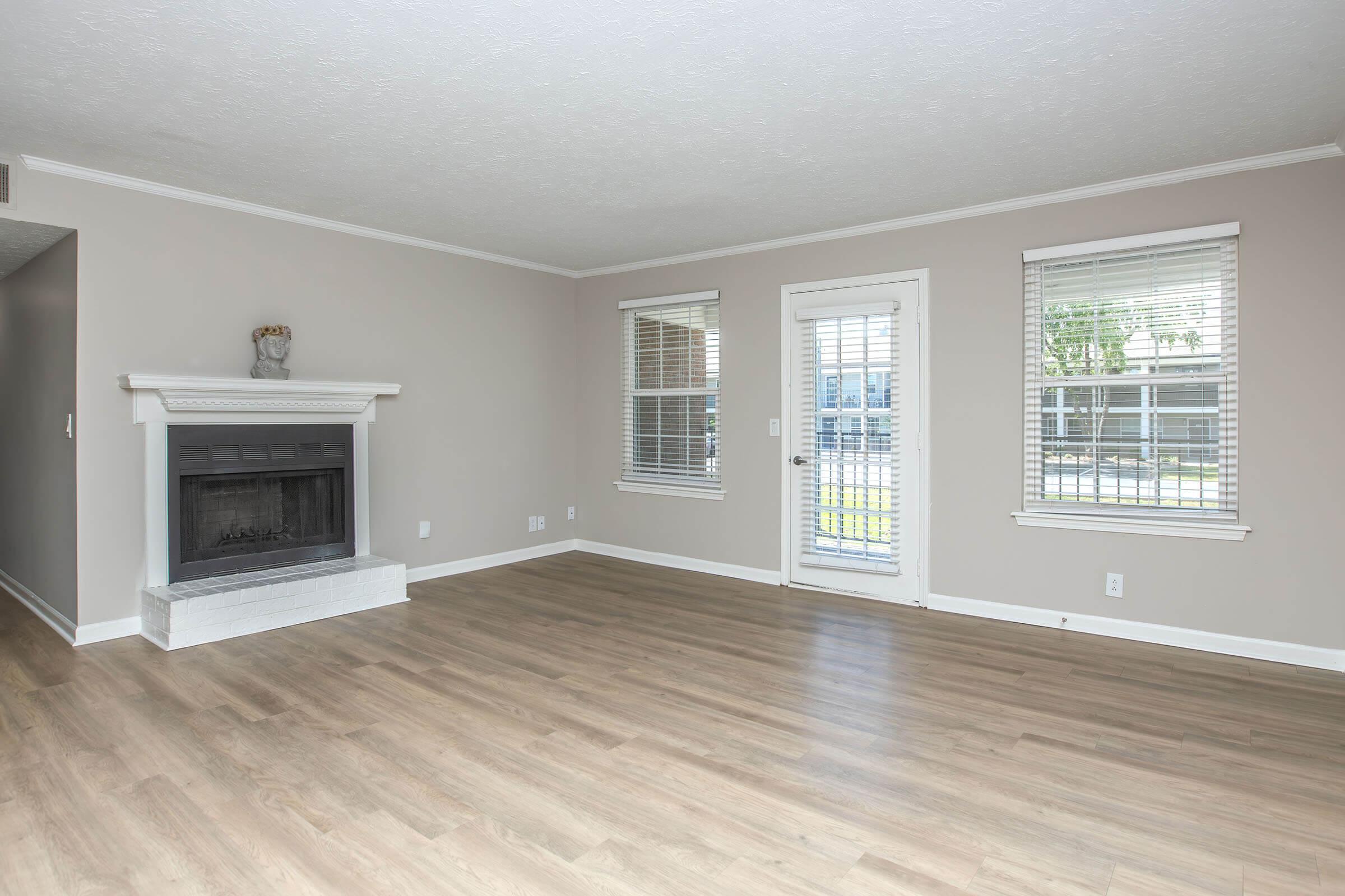 A spacious, unfurnished living room with light-colored walls and a large window. It features a white fireplace with a decorative item on top, and a sliding glass door leading to an outdoor area. The flooring is light wood, creating a bright and open atmosphere. Natural light floods the room, enhancing its inviting feel.
