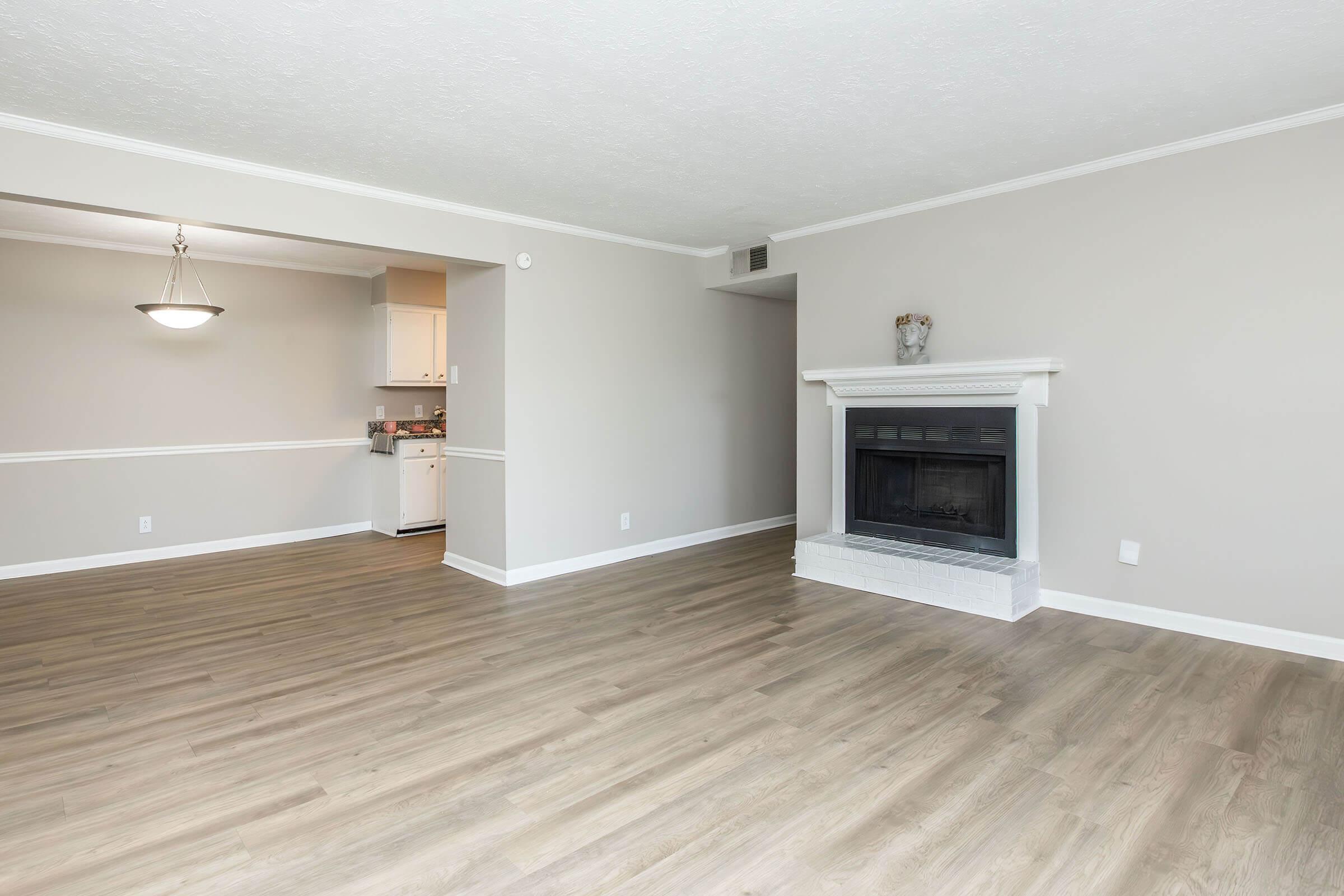 Empty living room with light brown laminate flooring, a white fireplace, and neutral-colored walls. A doorway leads to a kitchen area in the background, which appears bright and functional. A pendant light hangs from the ceiling, enhancing the open and spacious feel of the room.
