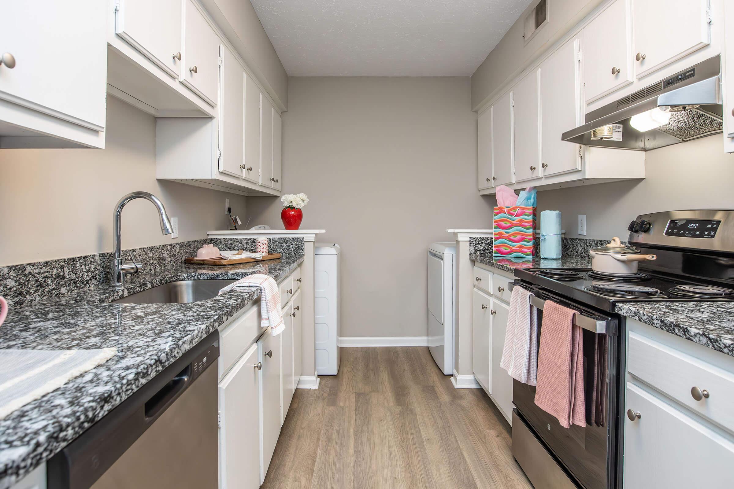 A modern kitchen featuring white cabinets and a granite countertop. The kitchen includes a sink, dishwasher, and stove with an oven. A washing machine and dryer are visible. The walls are painted a soft beige, and decorative items, including a vase and colorful bags, add a personal touch.