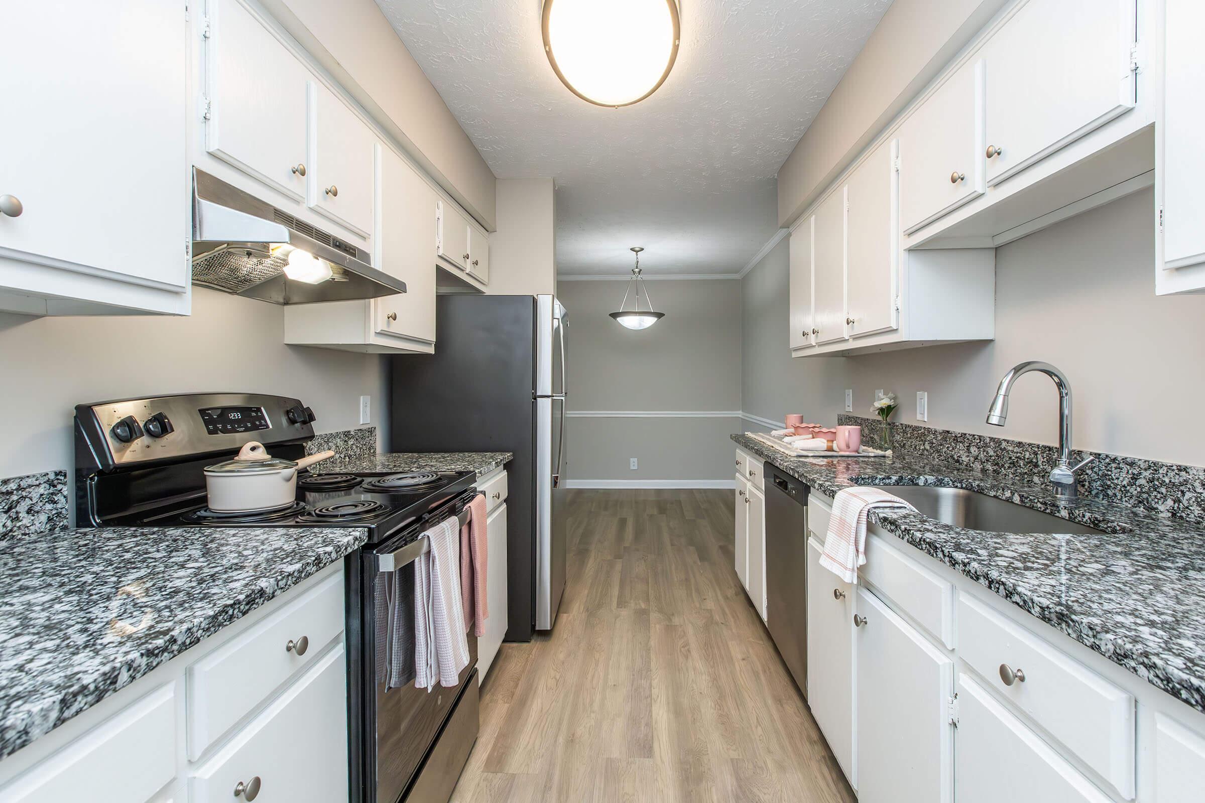 Modern kitchen featuring granite countertops, white cabinetry, stainless steel appliances, and a cozy dining area in the background. The space is well-lit with an overhead light fixture, and there are kitchen towels hanging from the oven.