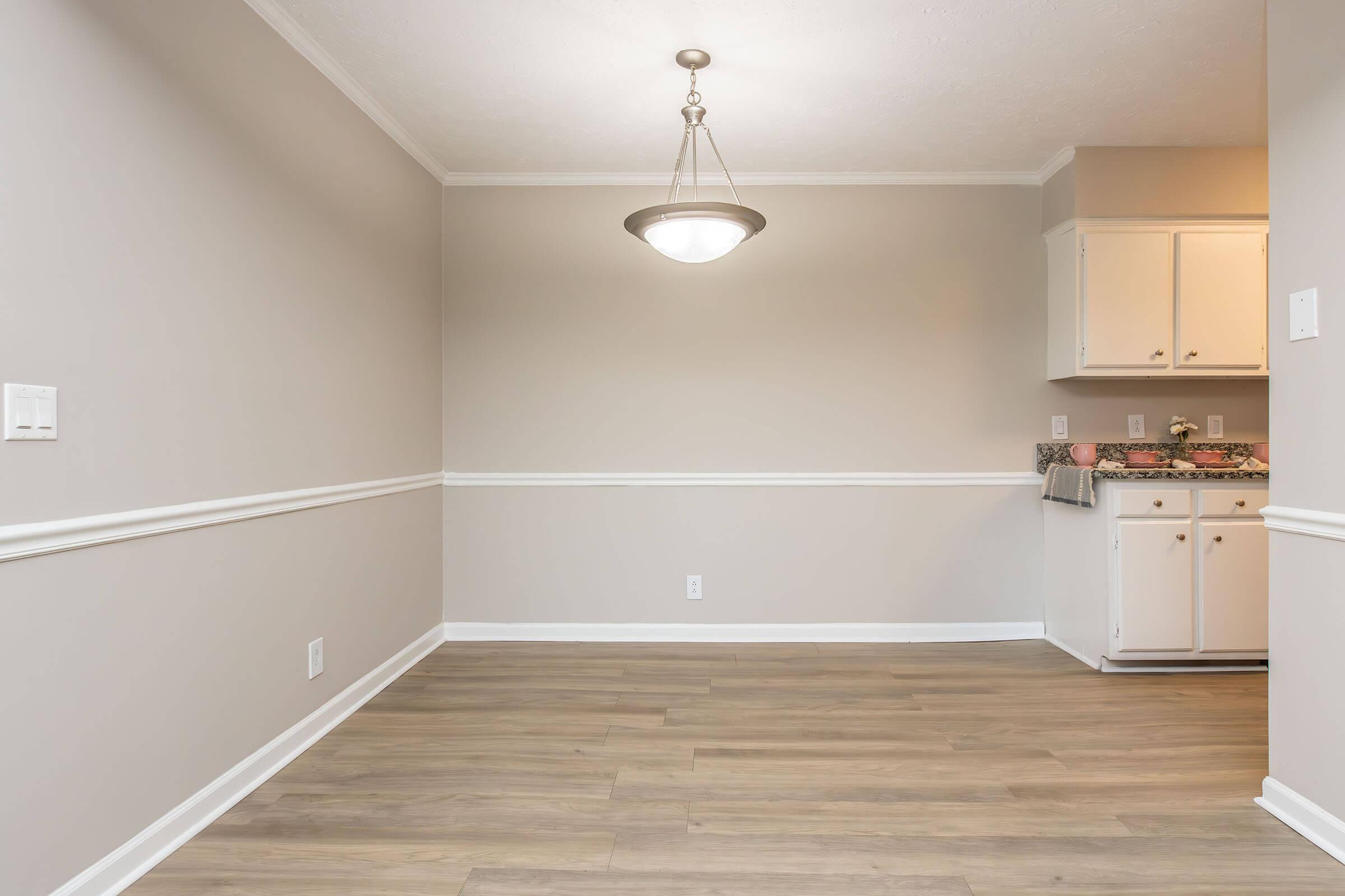 A spacious, empty room featuring light gray walls and a hardwood floor. There is a modern pendant light hanging from the ceiling, and a small kitchen area is visible in the background with white cabinets and a decorative countertop. A clean, minimalistic aesthetic is present throughout the space.