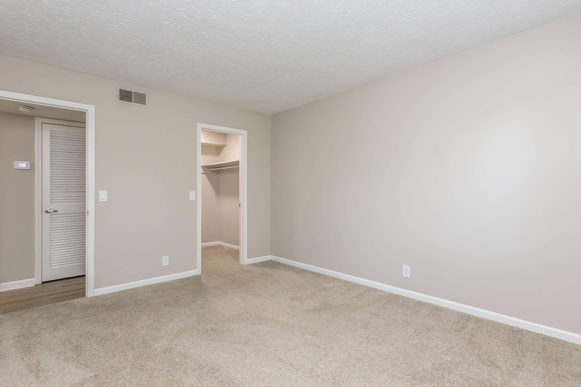 An empty room with light beige walls and carpet. A door on the left leads to a closet, while another door to the right opens to a hallway. The space is well-lit with natural light and features a simple, clean design, suitable for various furniture arrangements.