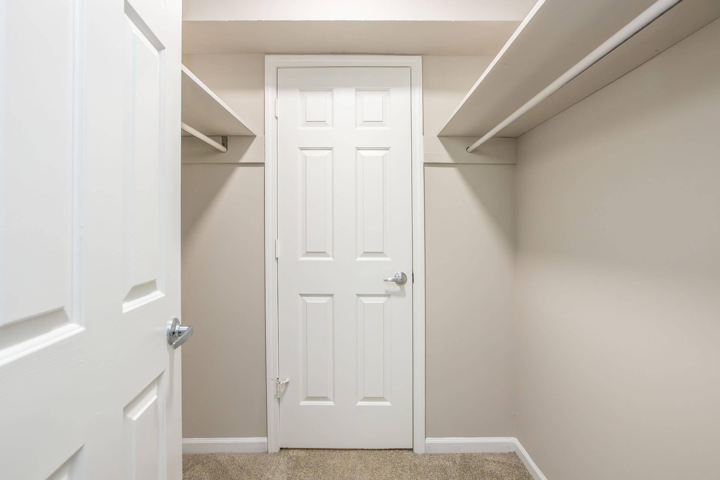 A small, empty closet featuring a plain beige wall, with two white shelving units on either side. A closed white door is visible at the end of the space, with a silver doorknob. The floor has a light brown carpet, giving the area a clean and simple appearance.
