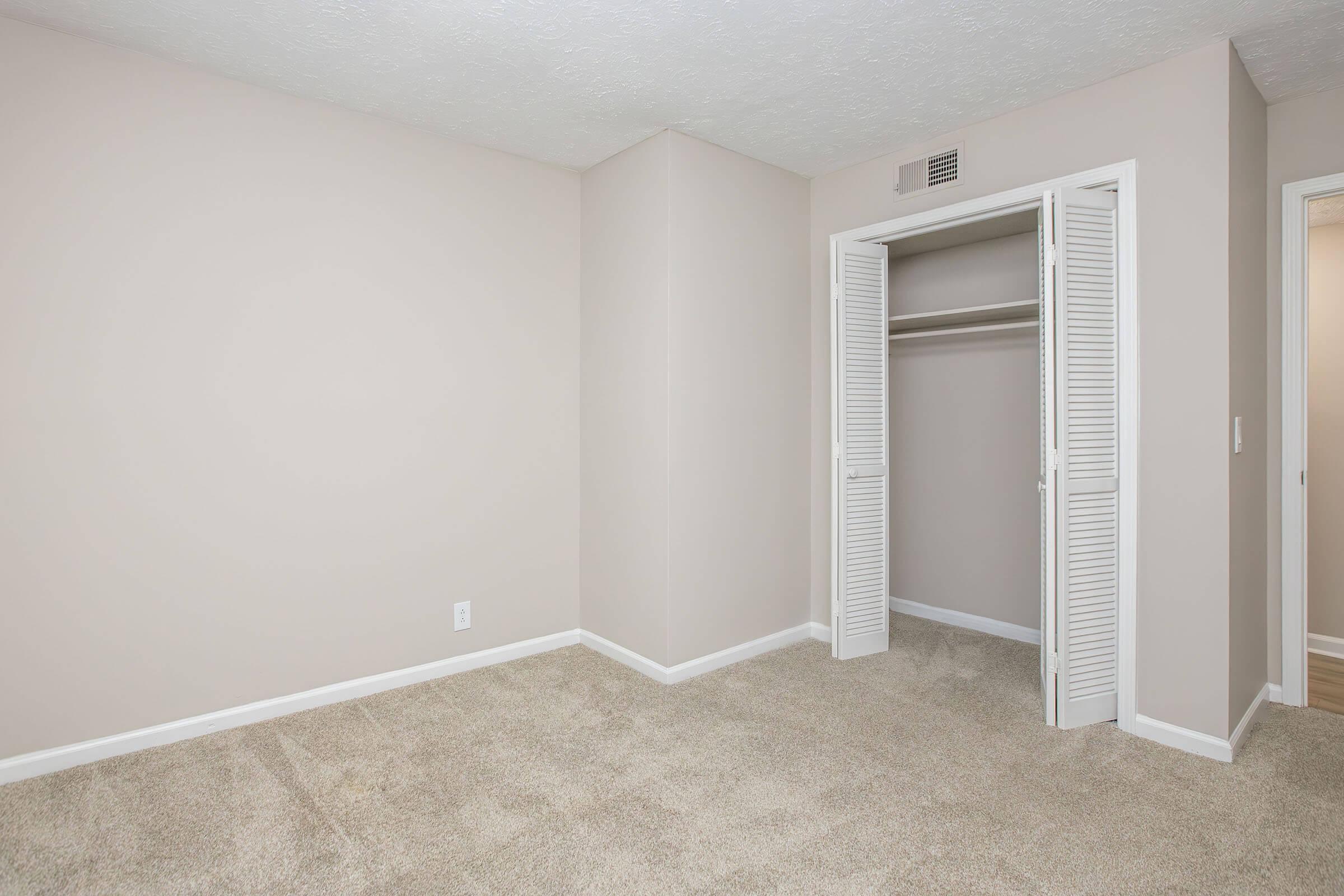 A clean, empty bedroom with light beige walls and carpet. It features a closet with white louvered doors. The room is well-lit and has a simple, spacious layout, creating a blank canvas for potential furnishings and decor.