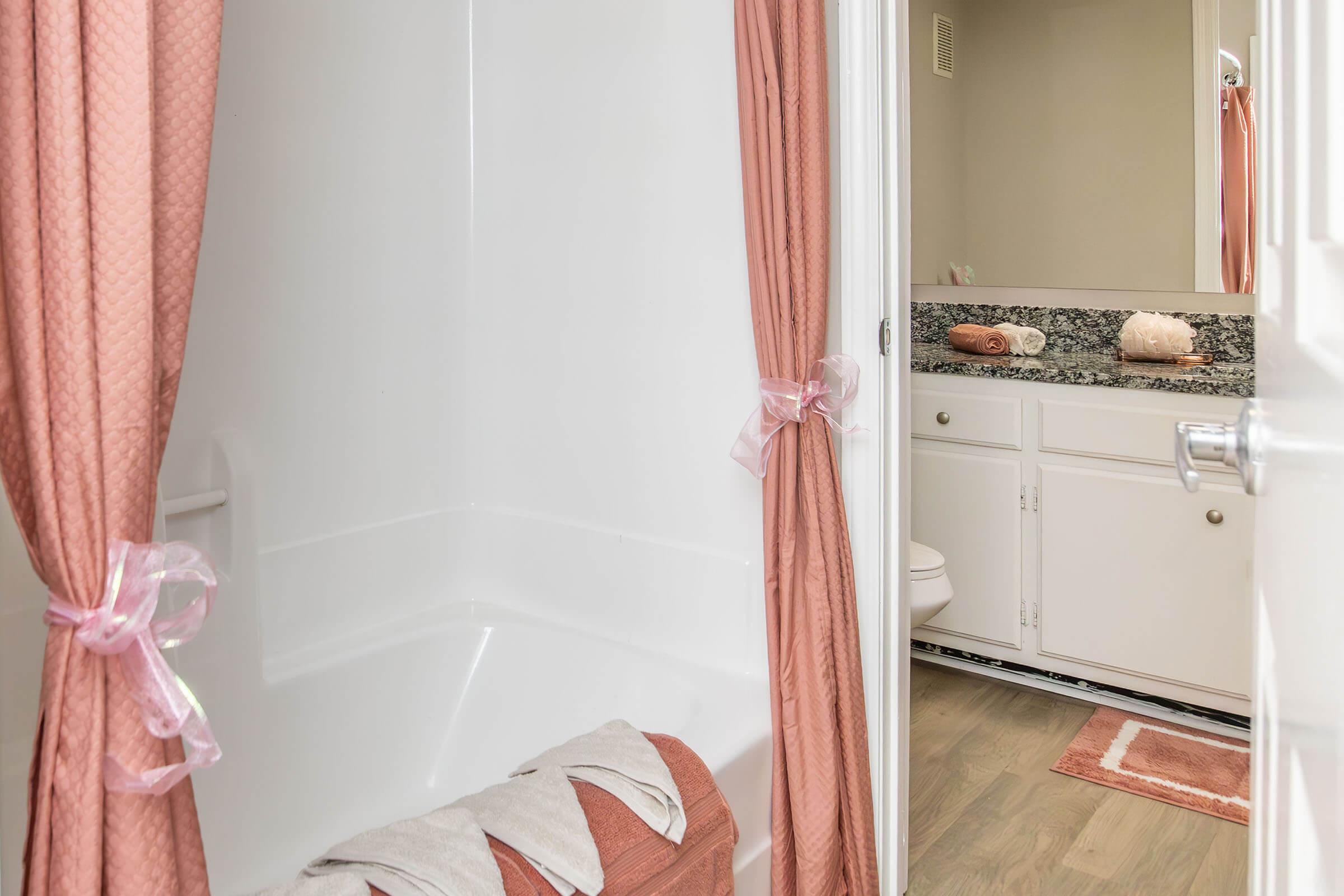 A clean bathroom with a shower and bathtub area, featuring peach-colored curtains tied with ribbons. The floor is covered with a small rug, and there's a granite countertop with neatly arranged toiletries. A white cabinet is visible in the background, adding a modern touch to the space.