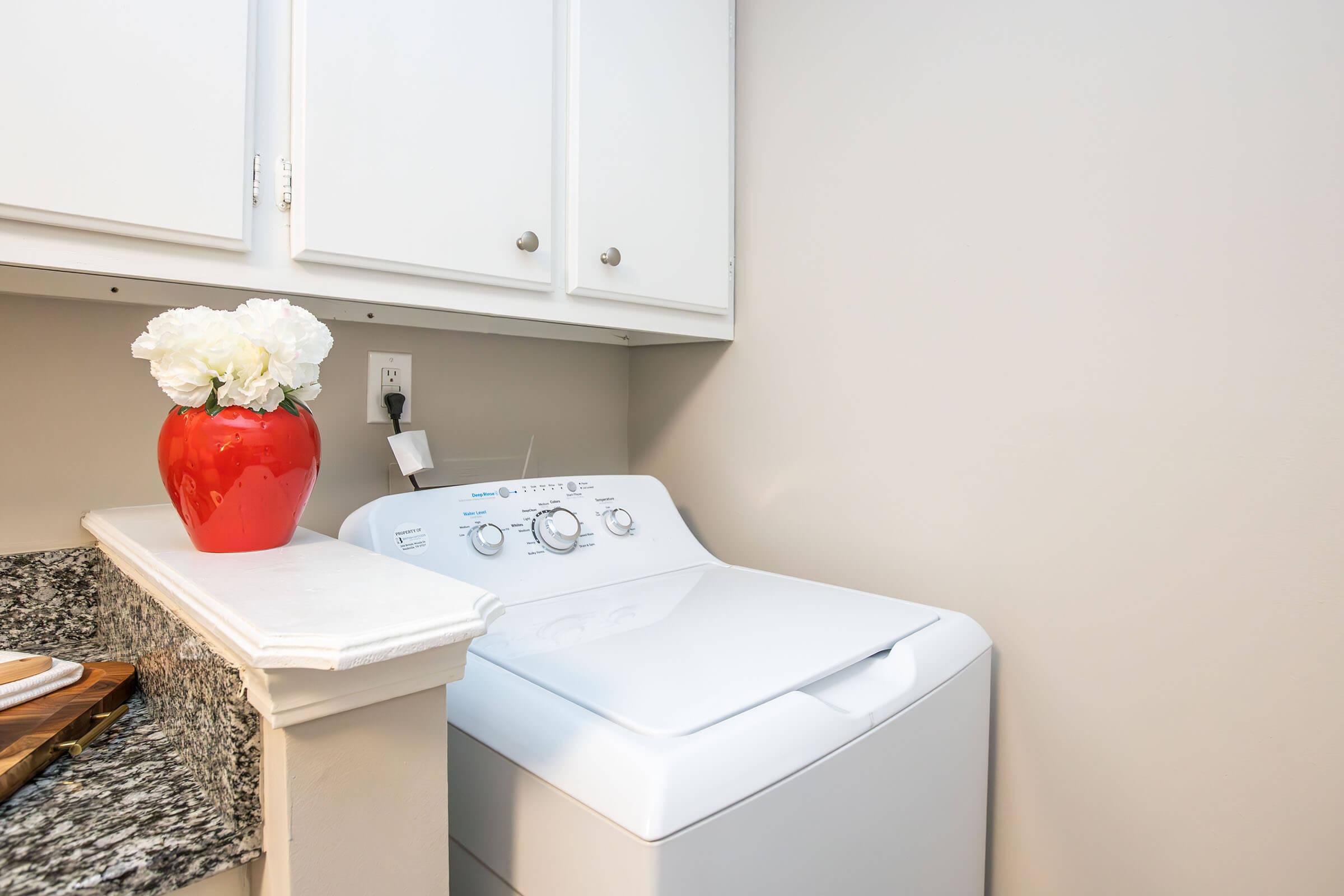 A laundry room featuring a white washing machine beside a granite countertop. On the countertop, there is a red vase with white flowers. The walls are painted in a neutral tone, and above the machine, there are white cabinets with silver handles.