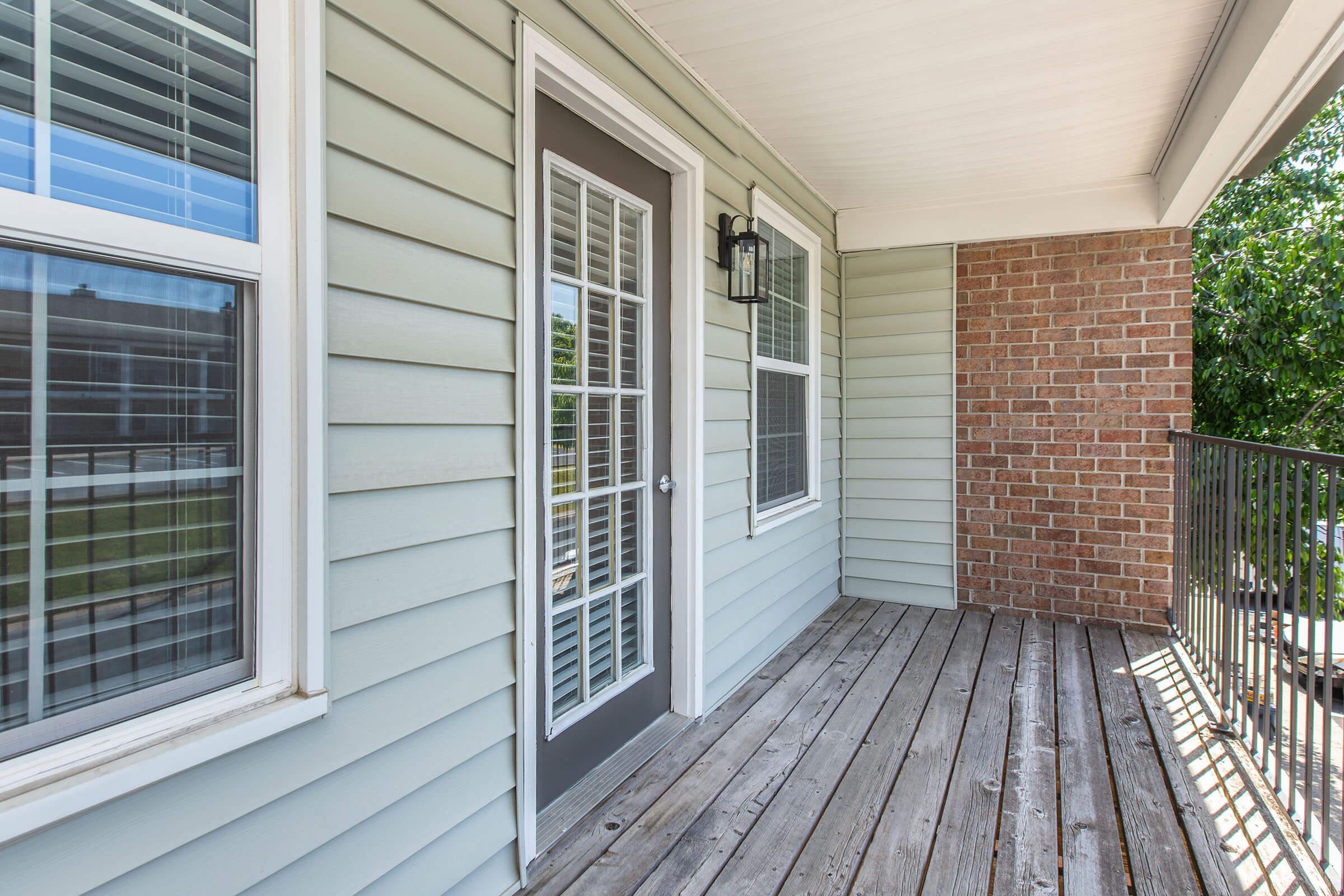 A well-lit porch area featuring a door with glass panels, surrounded by light gray wooden siding and a brick wall. The flooring is made of wooden planks, and there are large windows on either side of the door, providing natural light to the space.