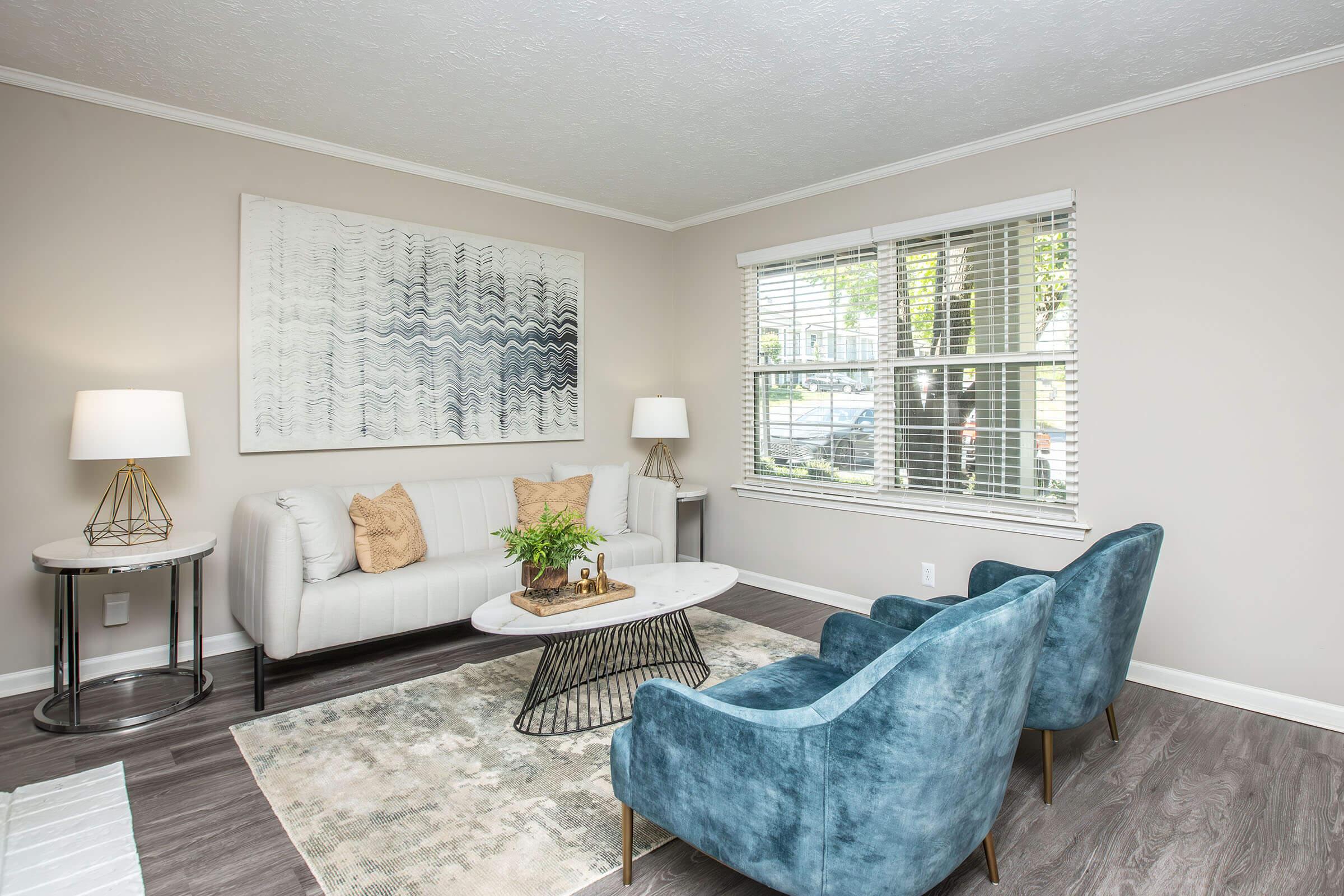 A modern living room featuring a light gray sofa with decorative pillows, two blue armchairs, a round coffee table with a black wire base, and a patterned area rug. Natural light filters in through a window with blinds, and there are two stylish lamps on side tables. A large wall art piece hangs above the sofa.