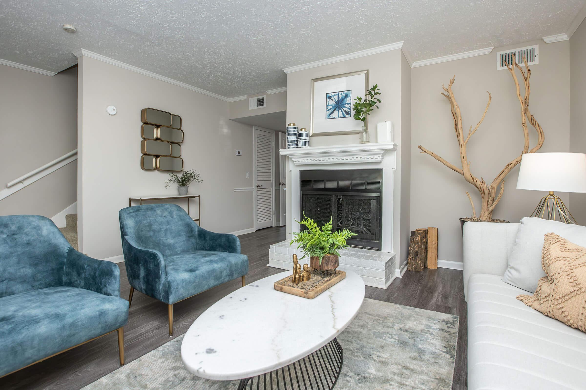 A stylish living room featuring a cozy white sofa with decorative pillows, two blue velvet chairs, a round marble coffee table, and a modern fireplace. A piece of art hangs above the fireplace, and there's a decorative branch display. The walls are painted in neutral tones, creating a warm and inviting atmosphere.