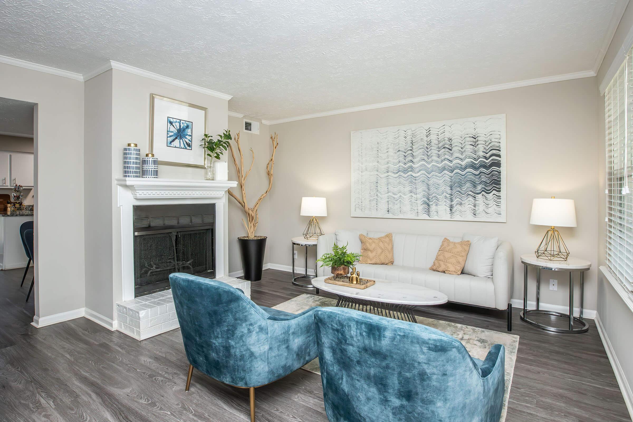 A modern living room featuring a light-colored sectional sofa with decorative pillows, two blue accent chairs, a round coffee table, and a fireplace. There is a large abstract wall art piece, potted plants, and two stylish lamps on either side of the sofa, creating a cozy and inviting atmosphere.
