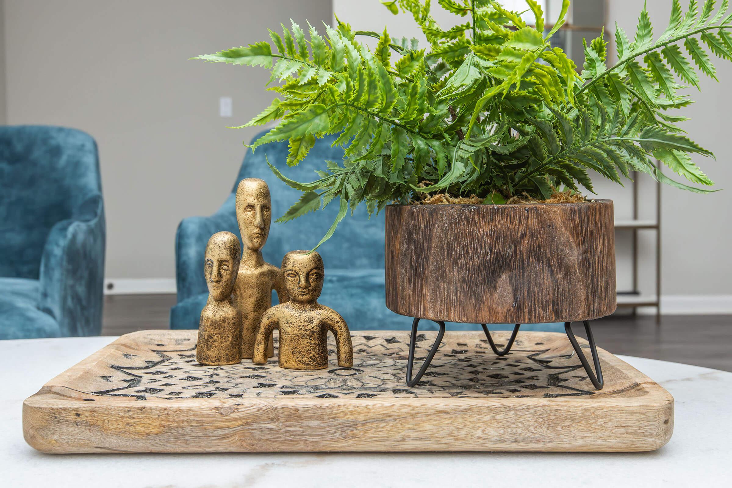 A wooden tray with intricate carvings holds a decorative plant in a round wooden pot with metal legs. Three small, gold-colored figurines of human-like faces are placed beside the plant, adding an artistic touch to the arrangement. The background features a soft blue chair and a neutral wall.