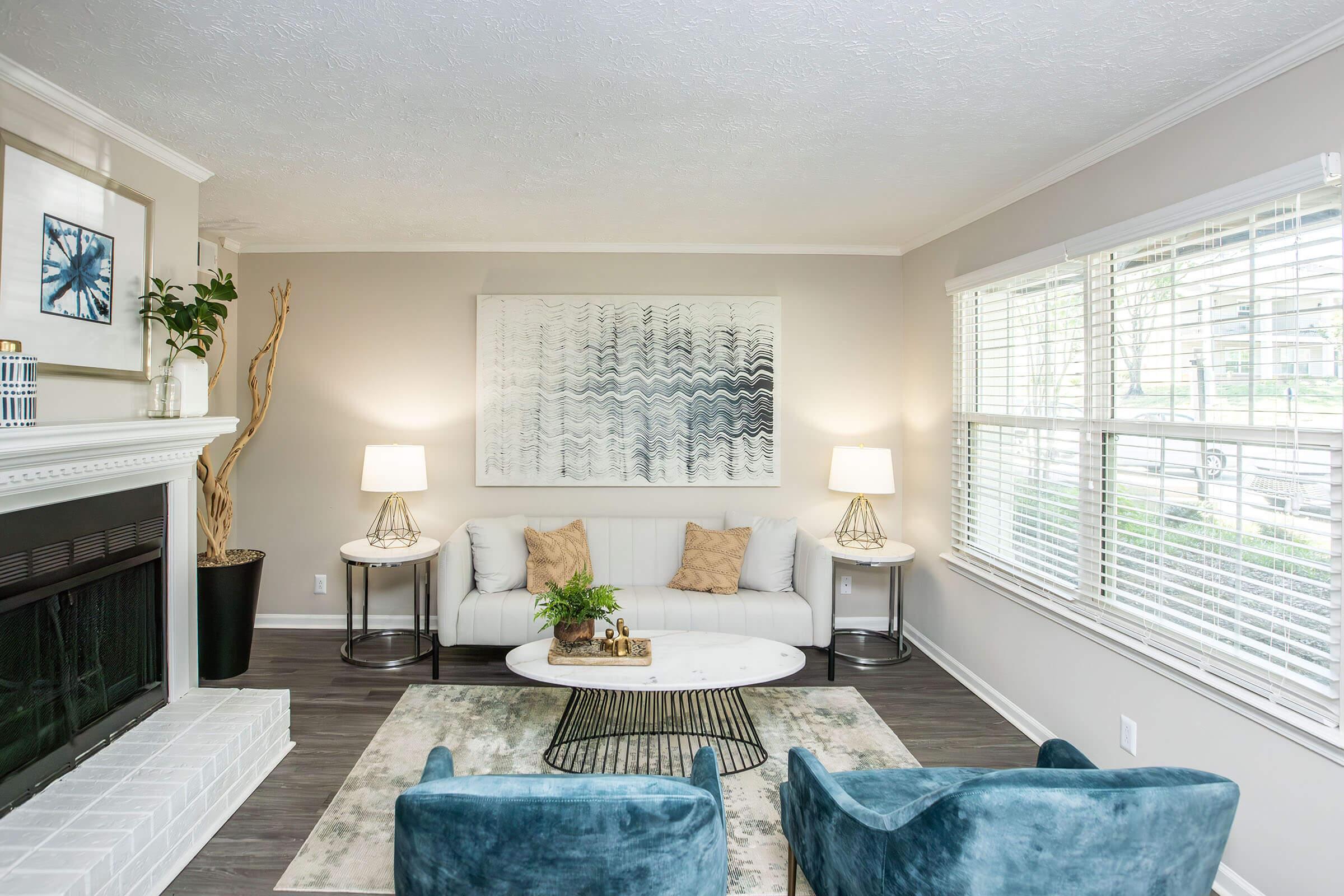 A modern living room featuring a white couch adorned with decorative pillows, two teal accent chairs, an oval coffee table on a patterned rug, and a fireplace. The walls are painted light gray, and large windows allow natural light to brighten the space. Two lamps and an abstract wall art piece add style.