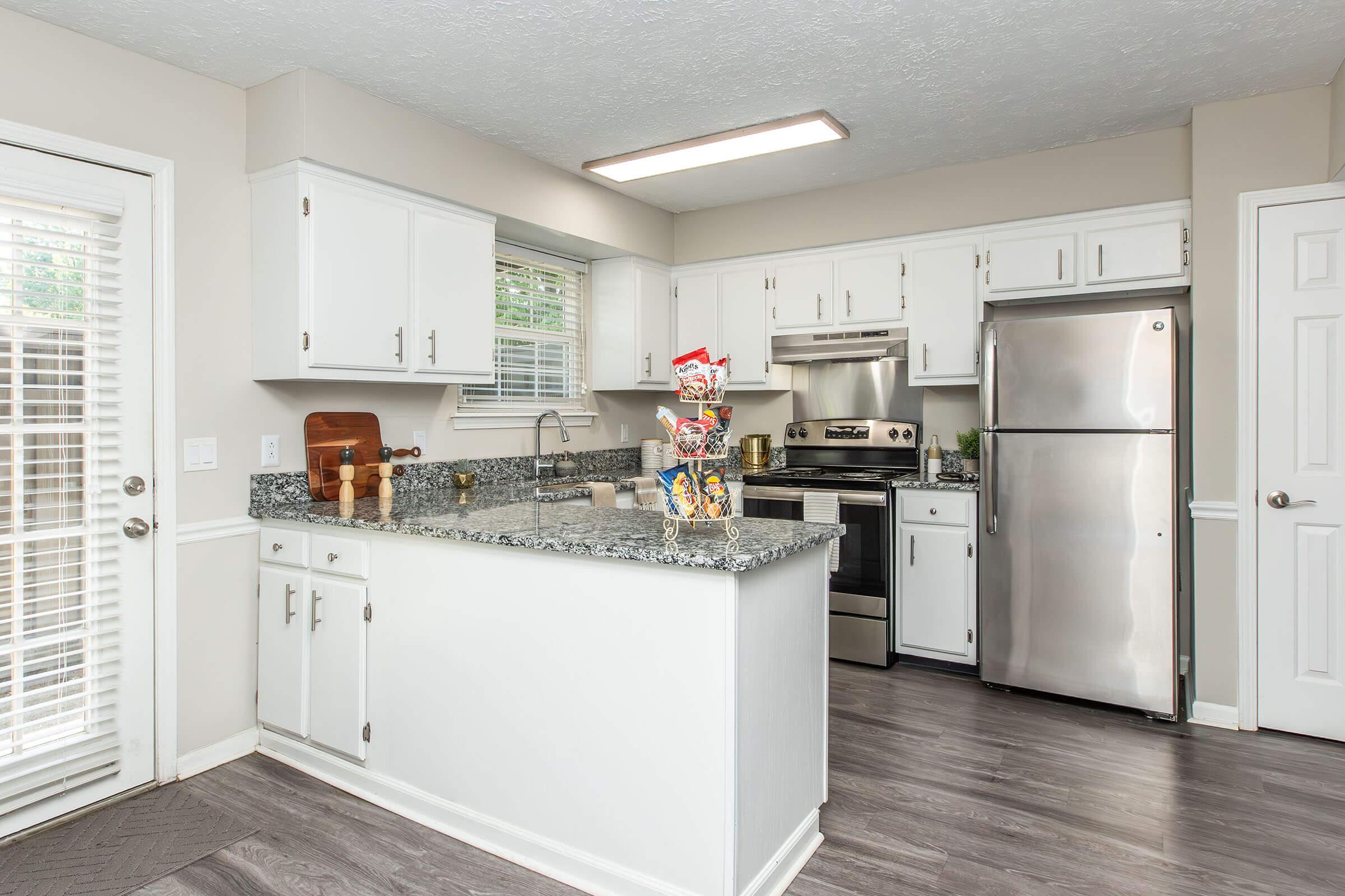 A modern kitchen featuring white cabinetry, stainless steel appliances, and a granite countertop. There is a silver refrigerator, an oven, and a sink with a window above it. A small decorative arrangement is on the counter, and the flooring is a warm wood pattern. Natural light comes from the sliding door.