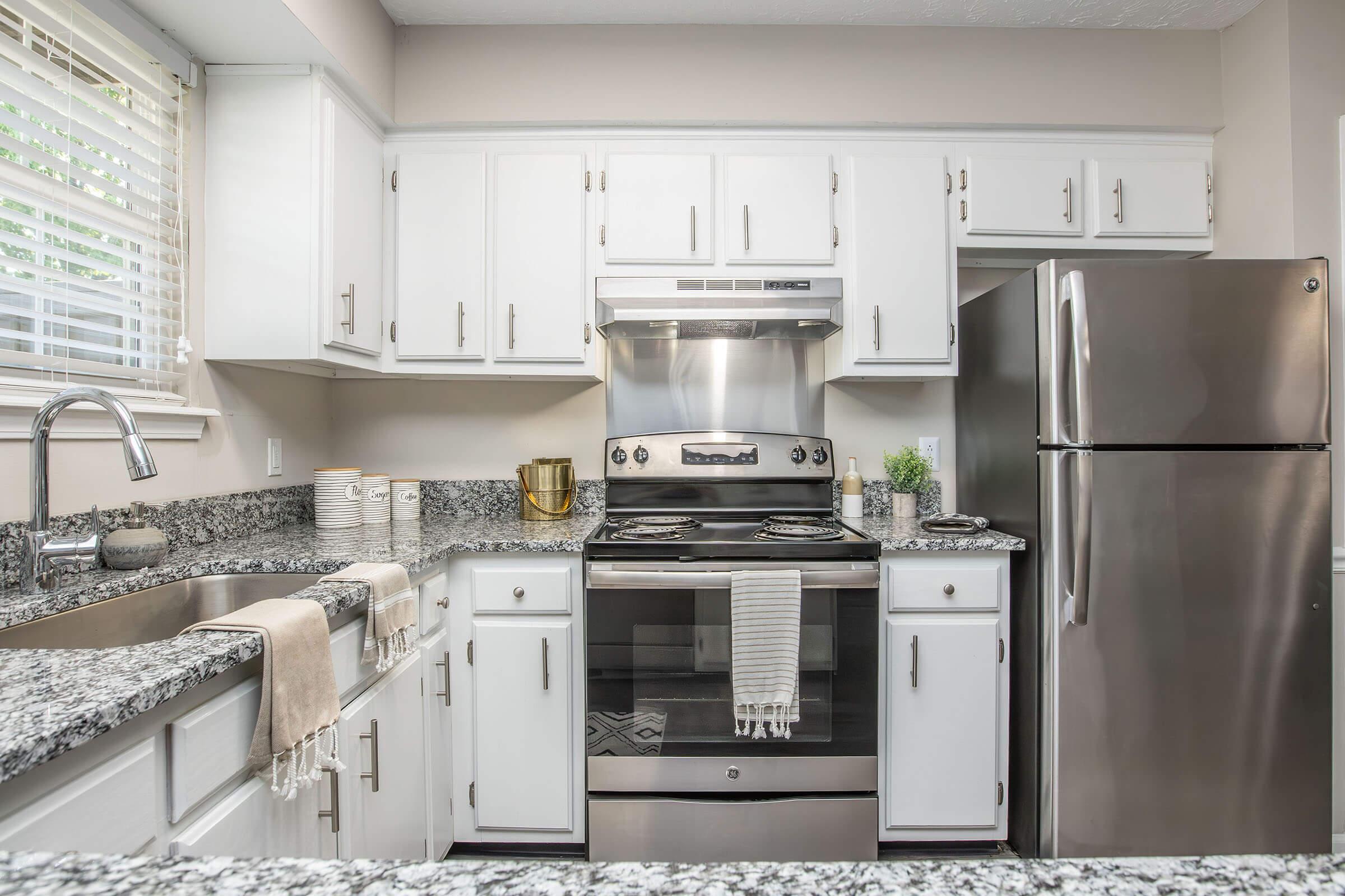 Modern kitchen featuring white cabinets, a stainless steel stove with a hood, and a silver refrigerator. The countertop is adorned with gray speckled granite. There's a window letting in natural light, and a bowl with decorative elements sits on the counter alongside neatly stacked plates. A towel hangs casually from the sink area.